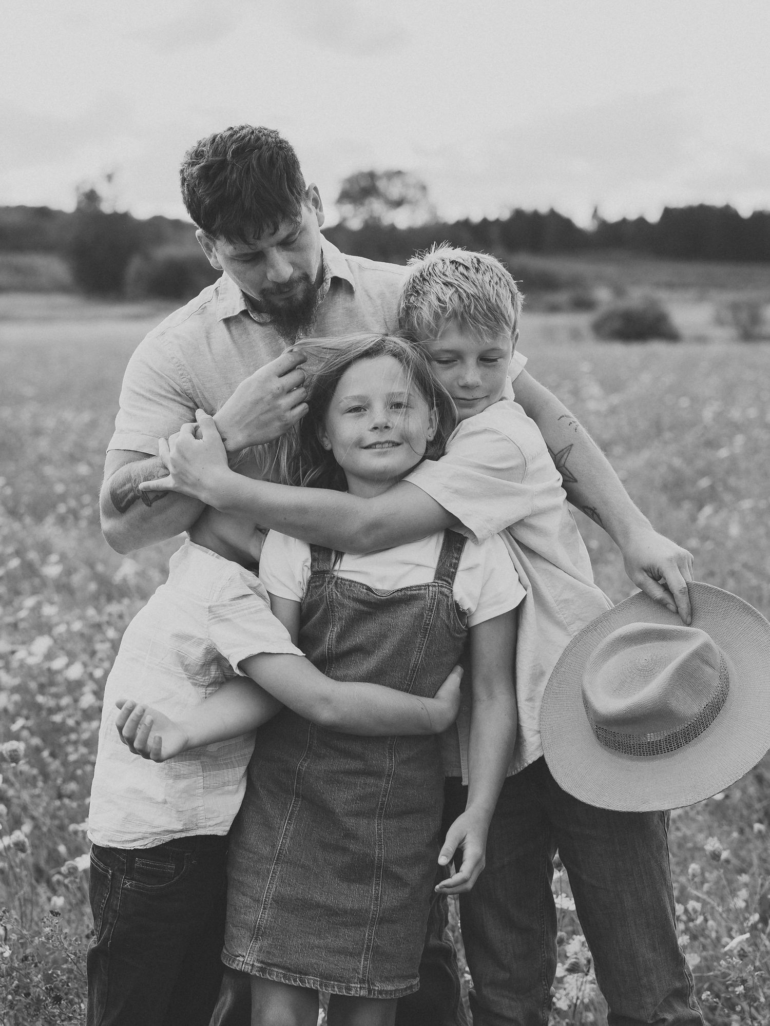 Black and white photo of a man hugging three children in a grassy field, one holding a wide-brimmed hat, in a warm family portrait.
