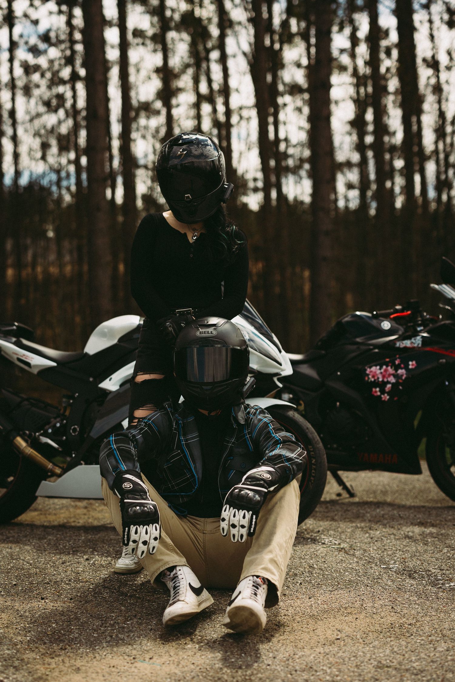 Two motorcyclists in helmets and gear pose near sport bikes in a forest setting.