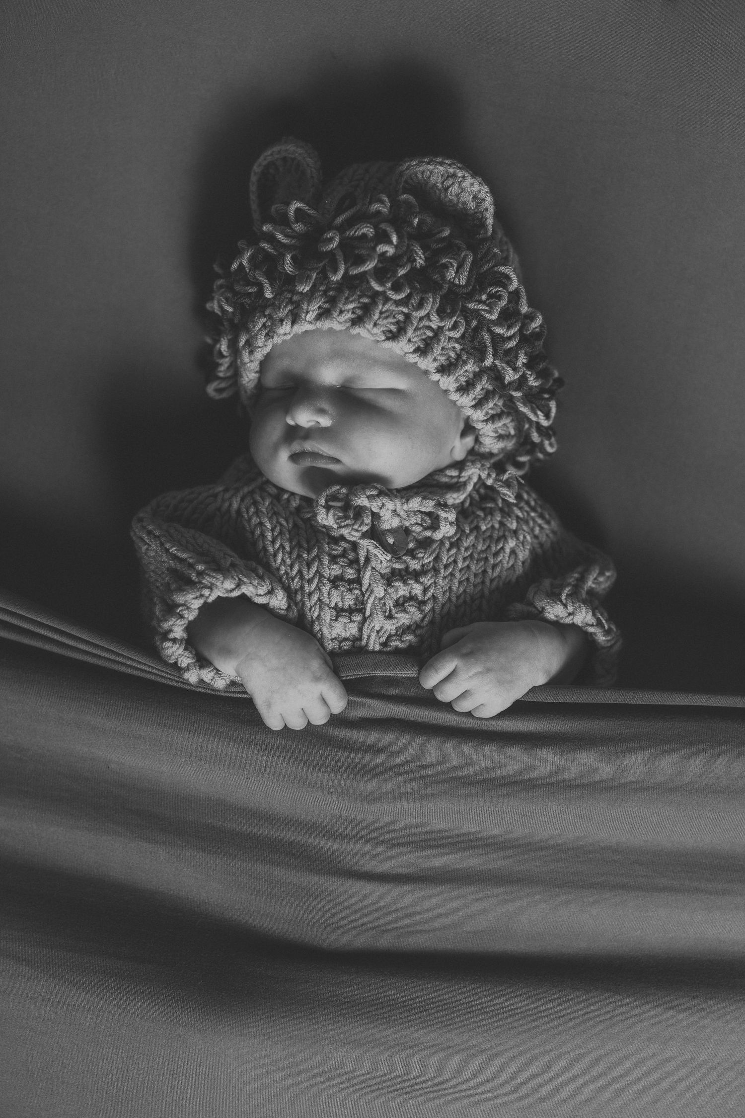 Black and white photo of newborn baby in knitted bear outfit and hat, sitting in soft fabric.