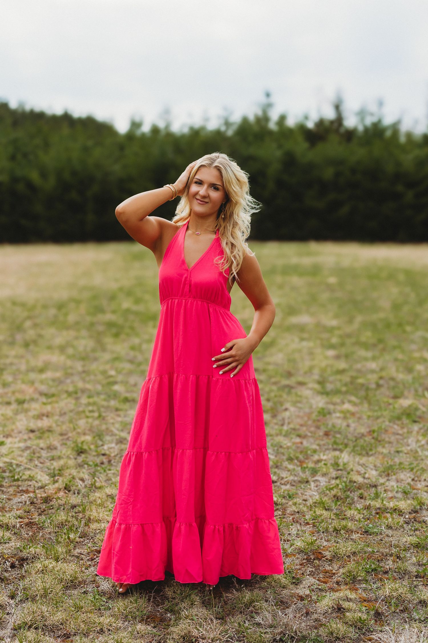 Blonde woman in a hot pink tiered maxi dress standing in a grassy field with evergreen trees behind her.