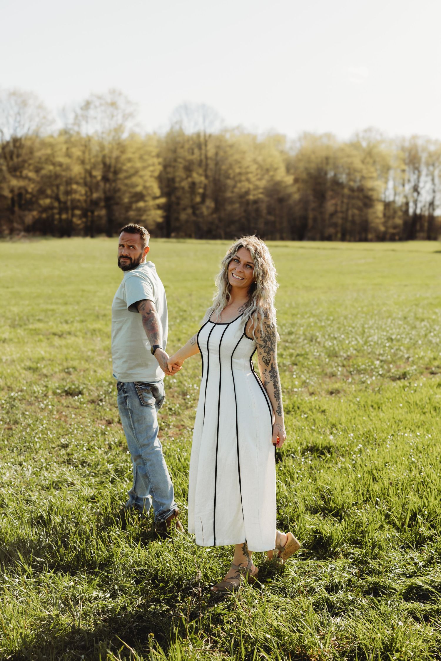 Tattooed couple holding hands in a green field, woman in white midi dress smiling, trees in background on sunny day.