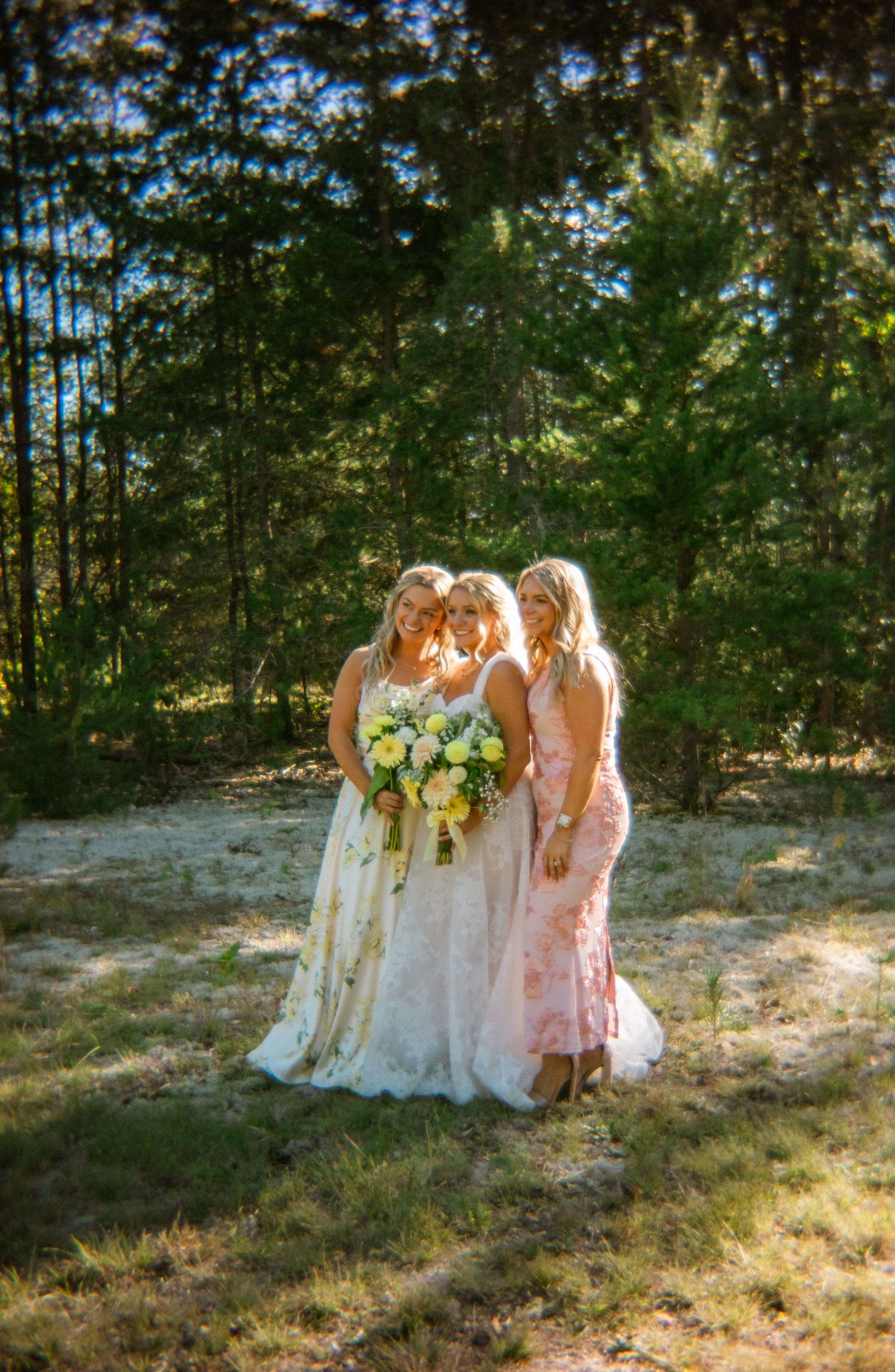 Three women in floral and pink bridesmaid dresses pose together holding a white and yellow bouquet in a sunlit forest clearing.