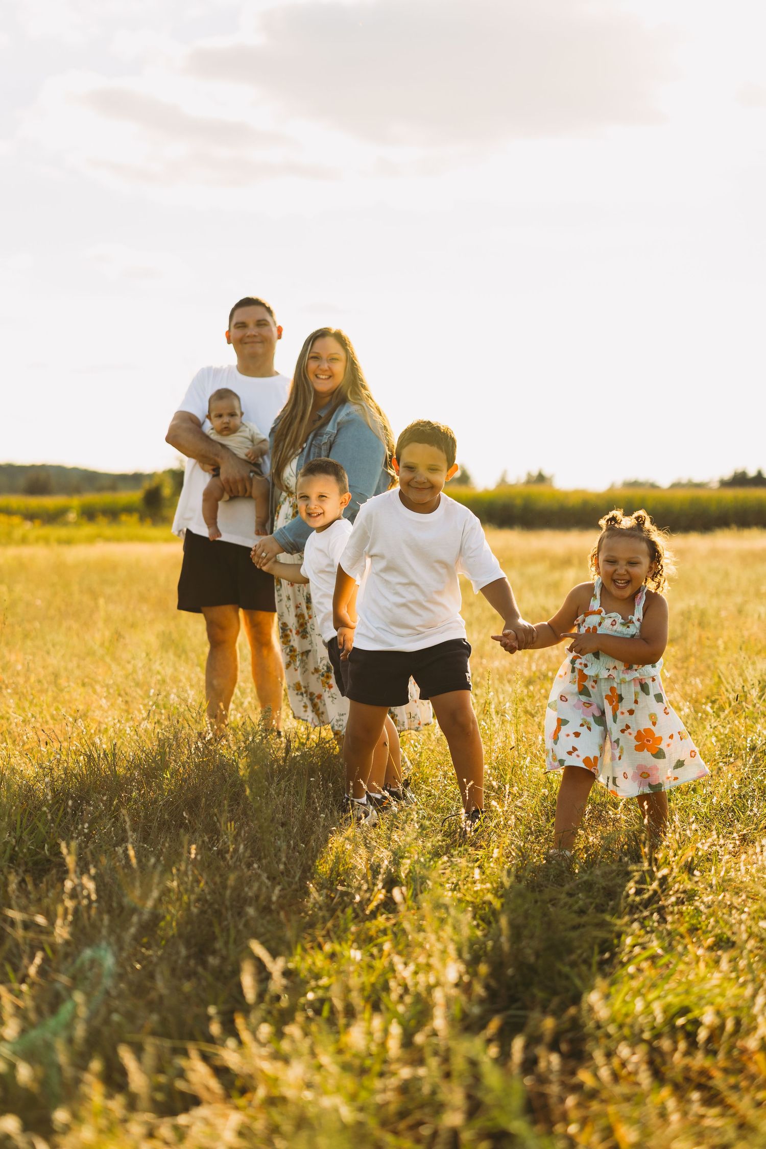 Smiling family of six playing together in a golden grassy field at sunset, with two parents holding a baby and three kids running.