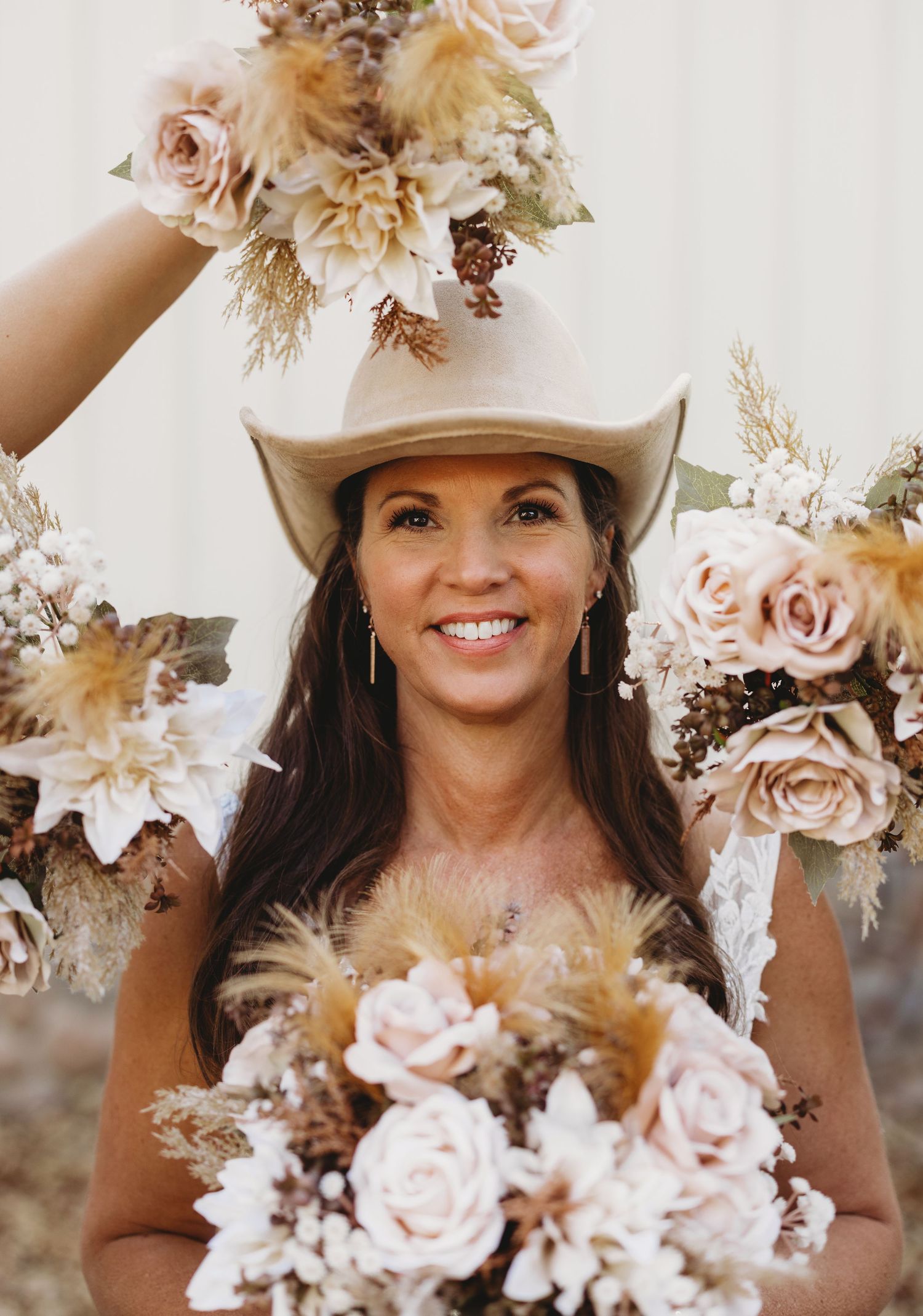 Smiling woman in a tan cowboy hat surrounded by boho bouquets of blush roses, pampas grass and dried flowers.