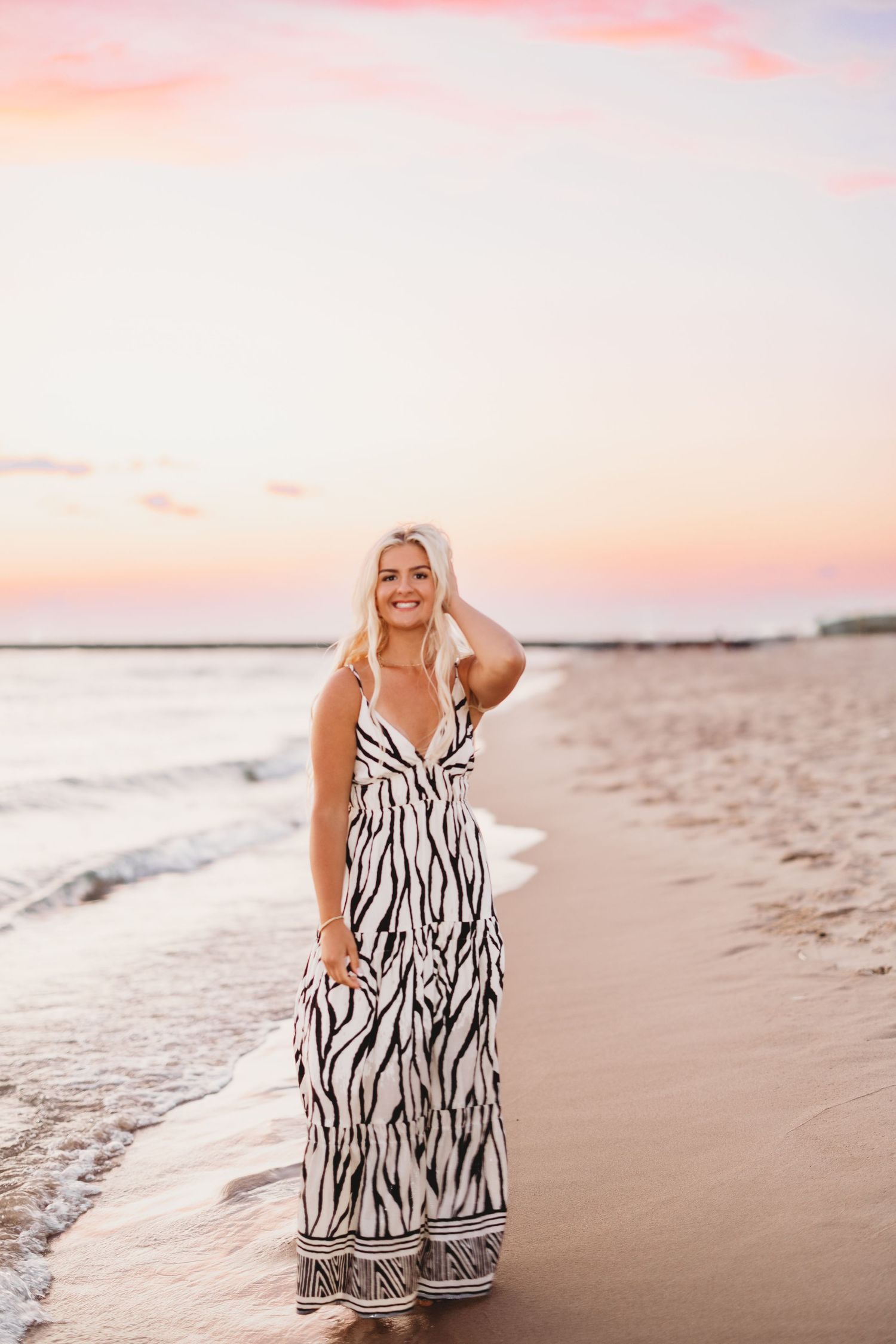 Smiling woman in a zebra print maxi dress stands at the water's edge on a beach beneath a pink sunset sky.