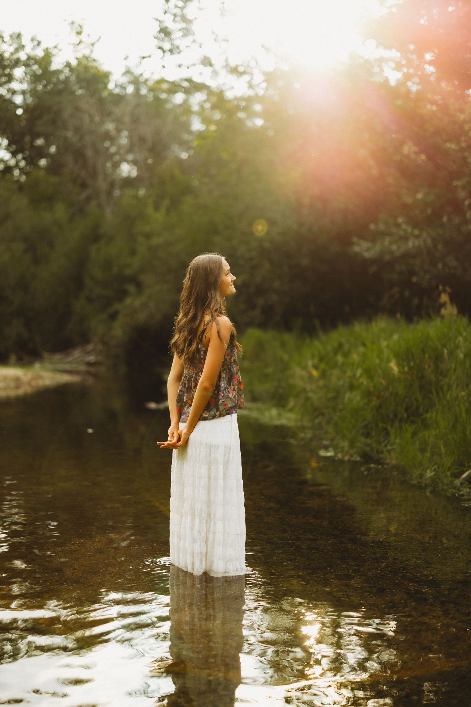 Woman in a white maxi skirt and floral top stands in shallow river water at golden hour, with sunlight flaring through the trees.