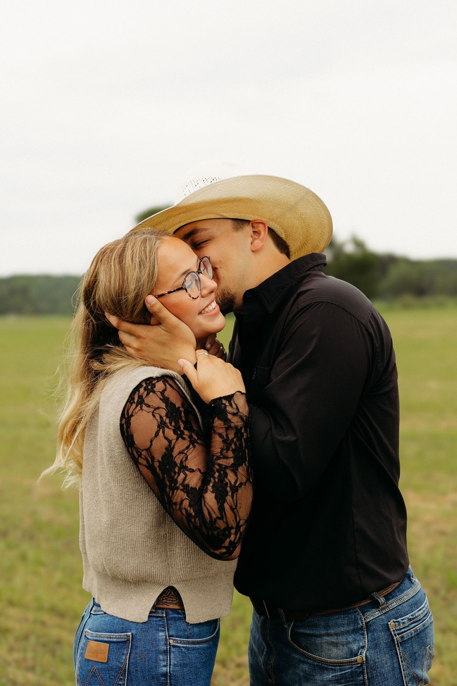 Couple sharing a romantic kiss in a green field, woman in lace top and vest, man in black shirt and cowboy hat.