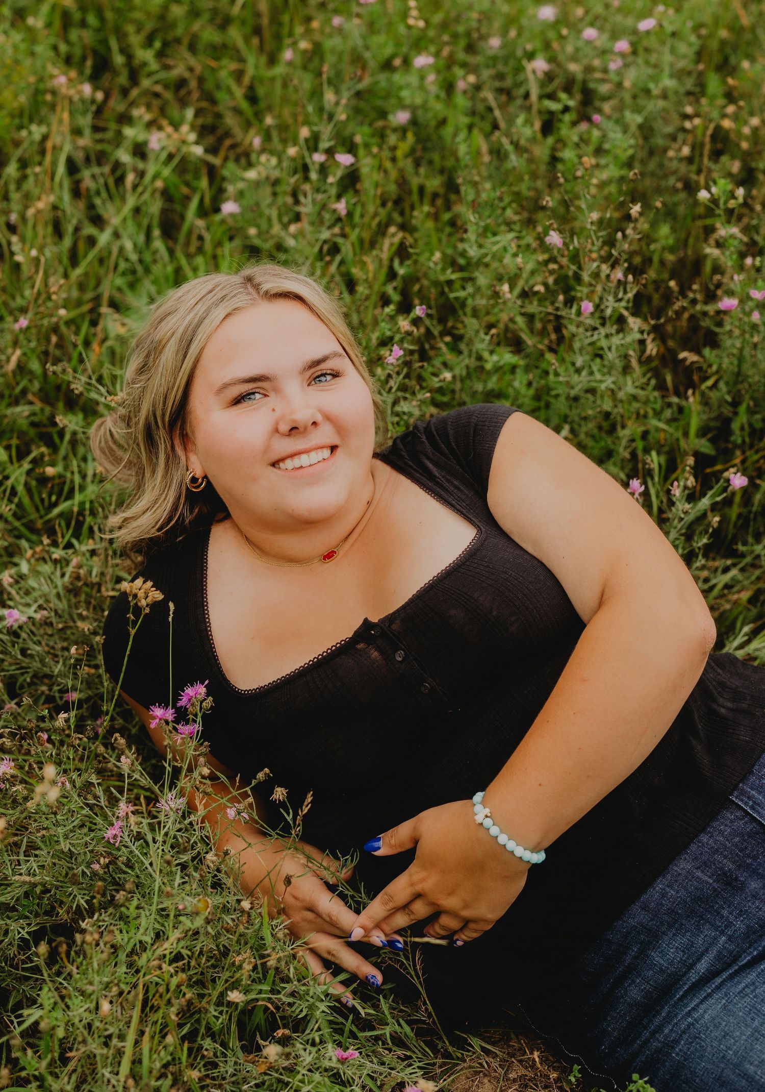 Young woman in black top sitting in a wildflower meadow, smiling and holding flowers on a sunny day.