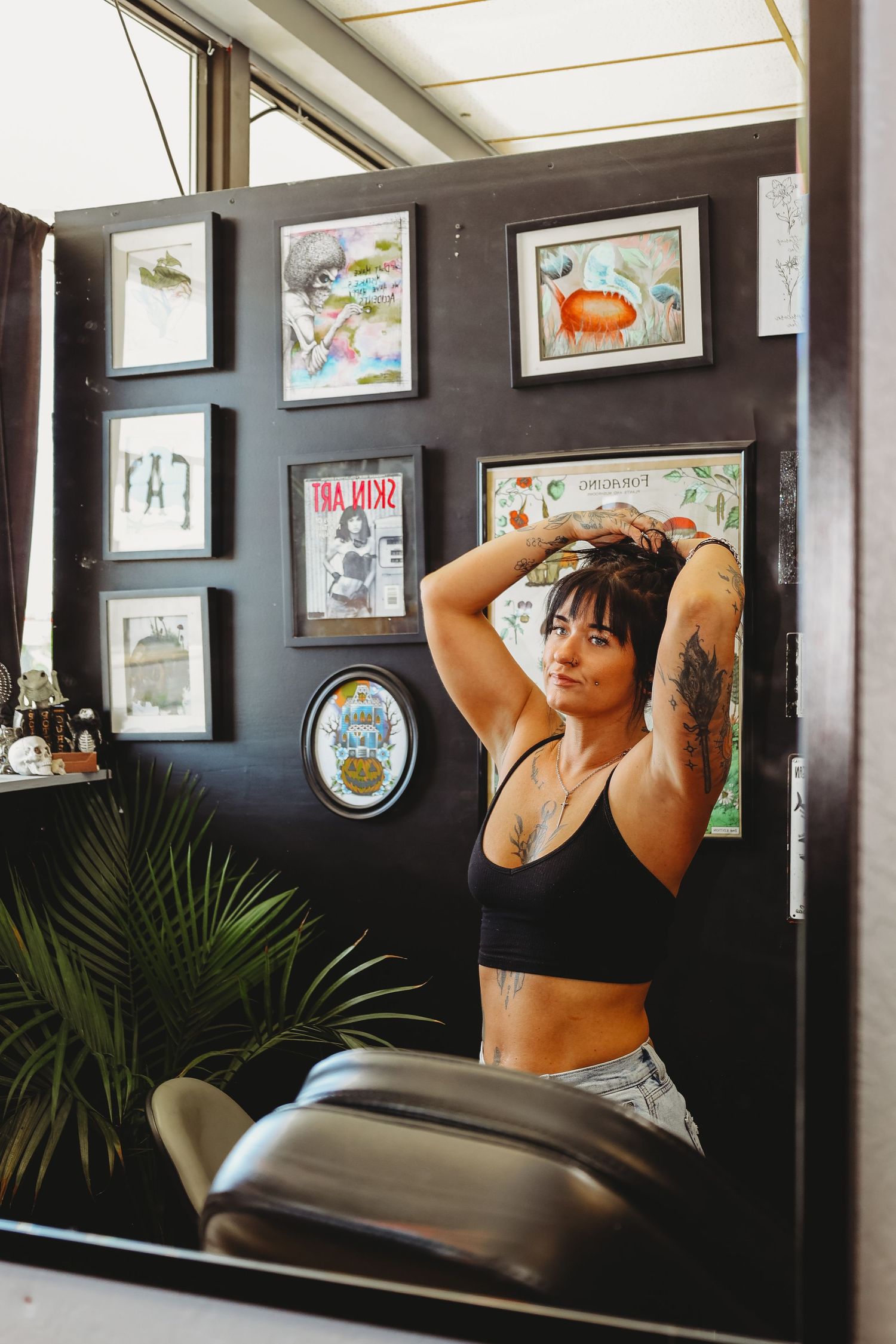 Tattooed woman in black crop top poses with hands behind head in a tattoo studio with framed artwork on dark walls.