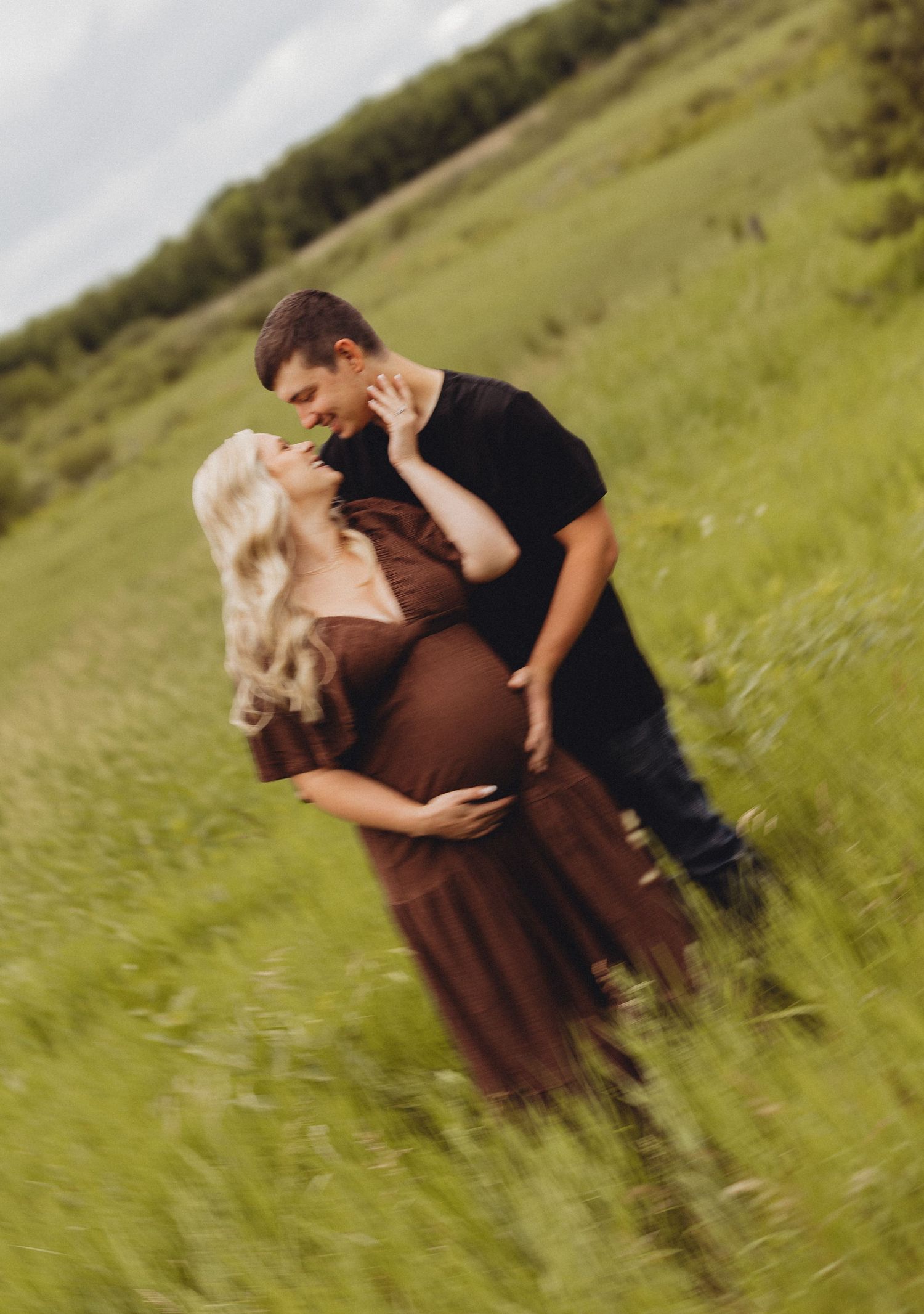 A pregnant woman in a brown dress embraces her partner in a lush green field during a romantic maternity photo shoot.