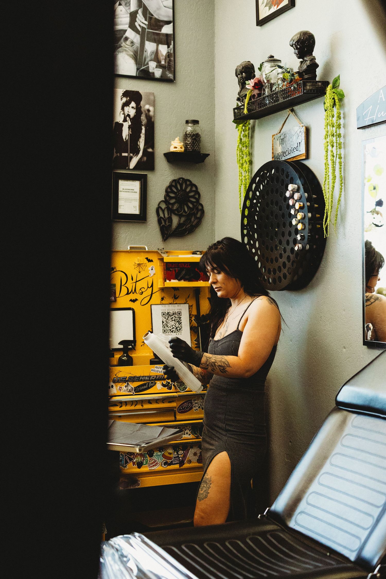 Female tattoo artist with tattoos reviews design in a decorated studio with yellow toolbox and framed artwork.
