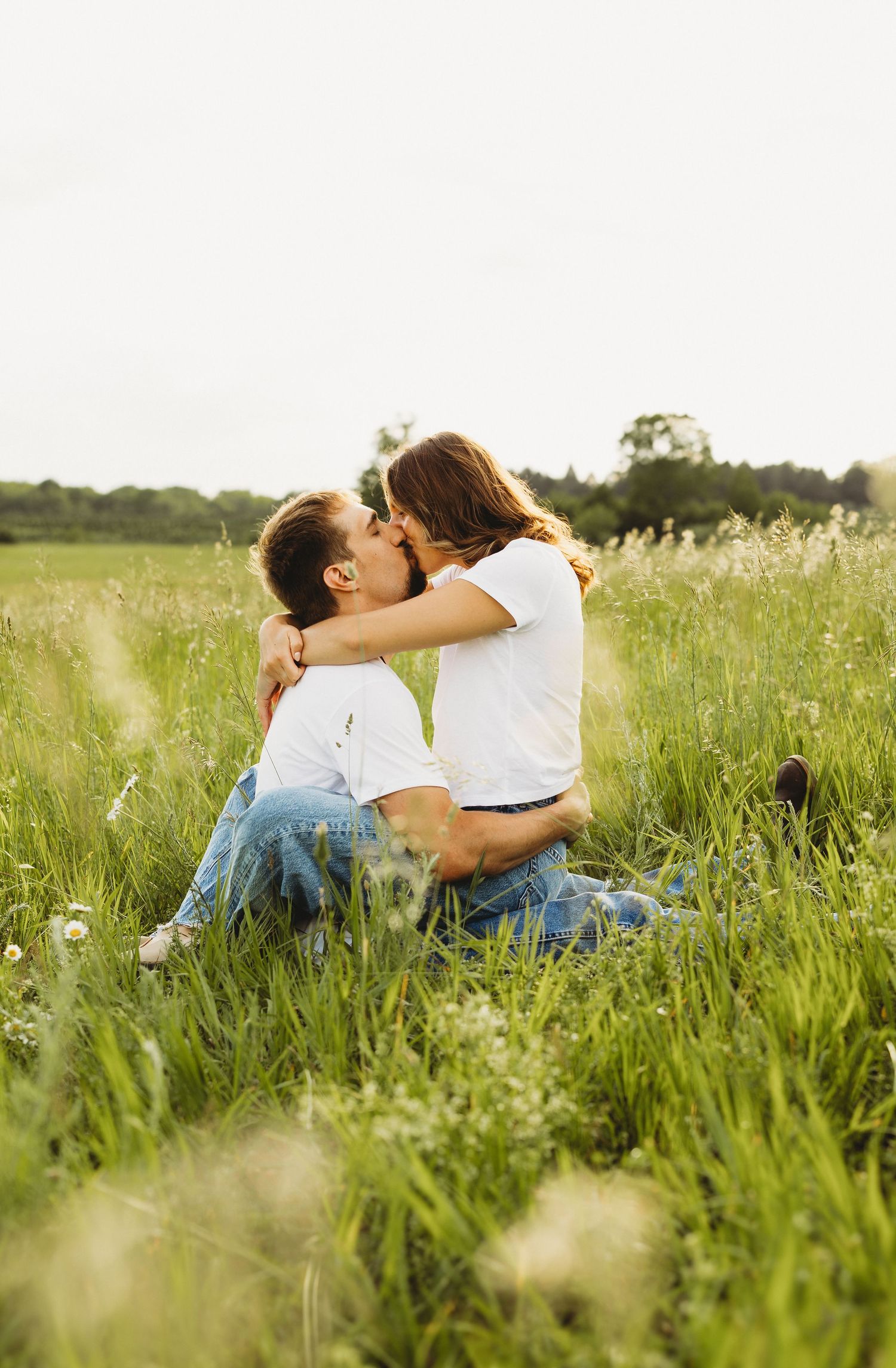 A couple sharing a romantic kiss while sitting in a lush green meadow field on a warm summer day.