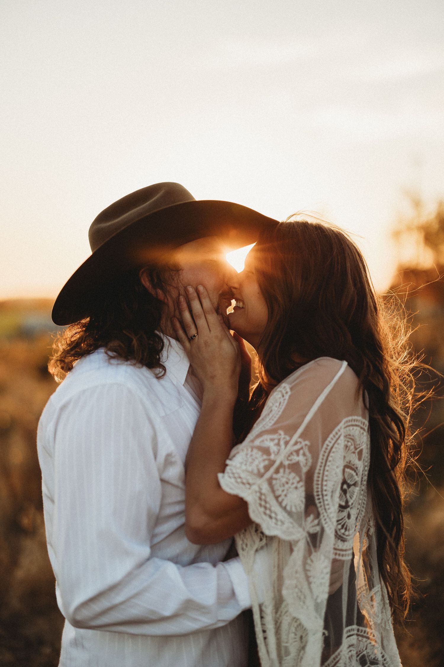 A romantic couple embraces in a white dress and cowboy hat during golden hour sunset in a rustic outdoor setting.