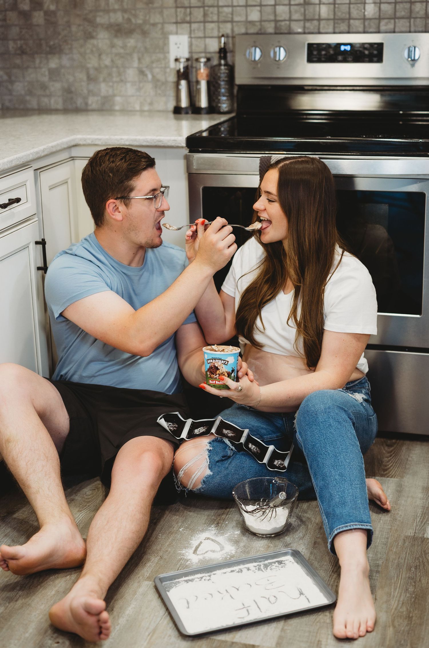 Couple sitting on kitchen floor sharing ice cream, laughing together near stove with baking sheet nearby.