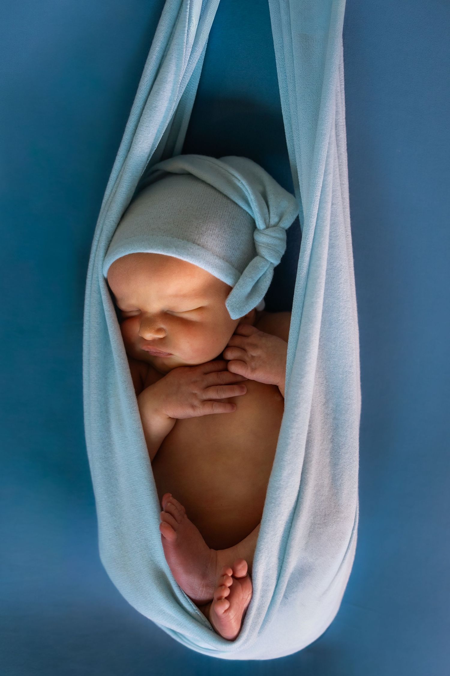 Newborn baby sleeping peacefully in a light blue fabric hammock wrap, wearing a soft knotted hat.