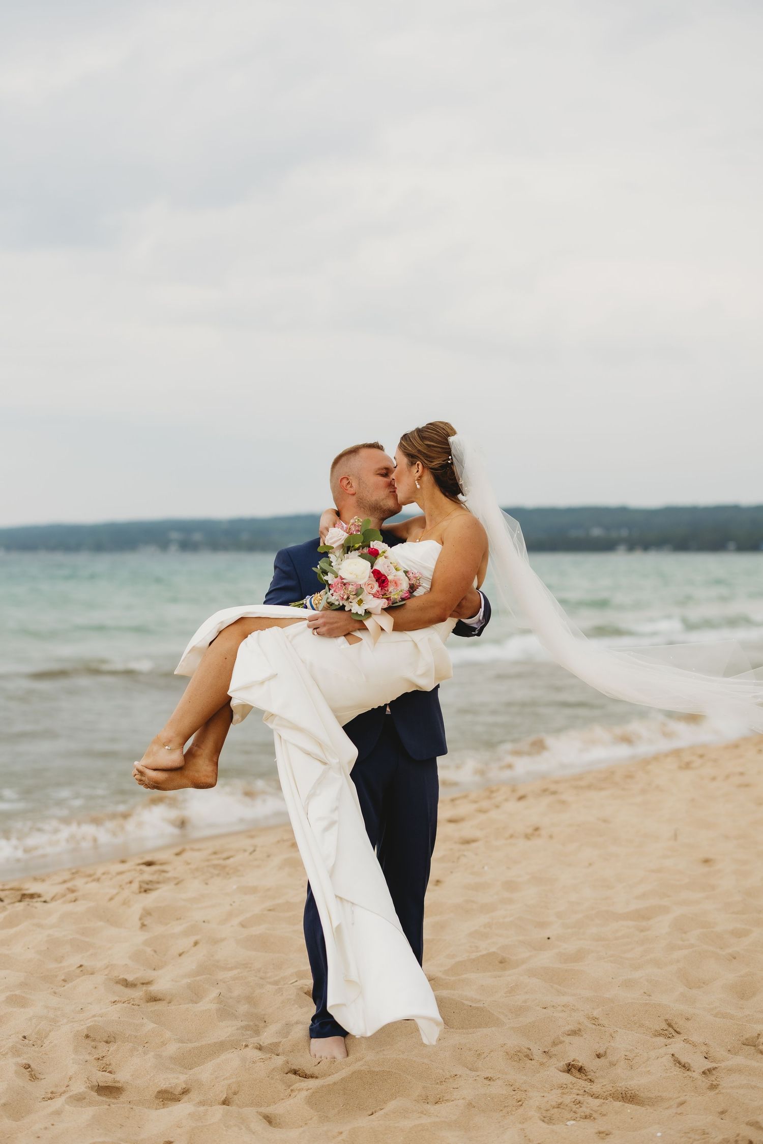 Groom carries bride in his arms while kissing on a sandy beach, her veil flowing in the wind with ocean backdrop.