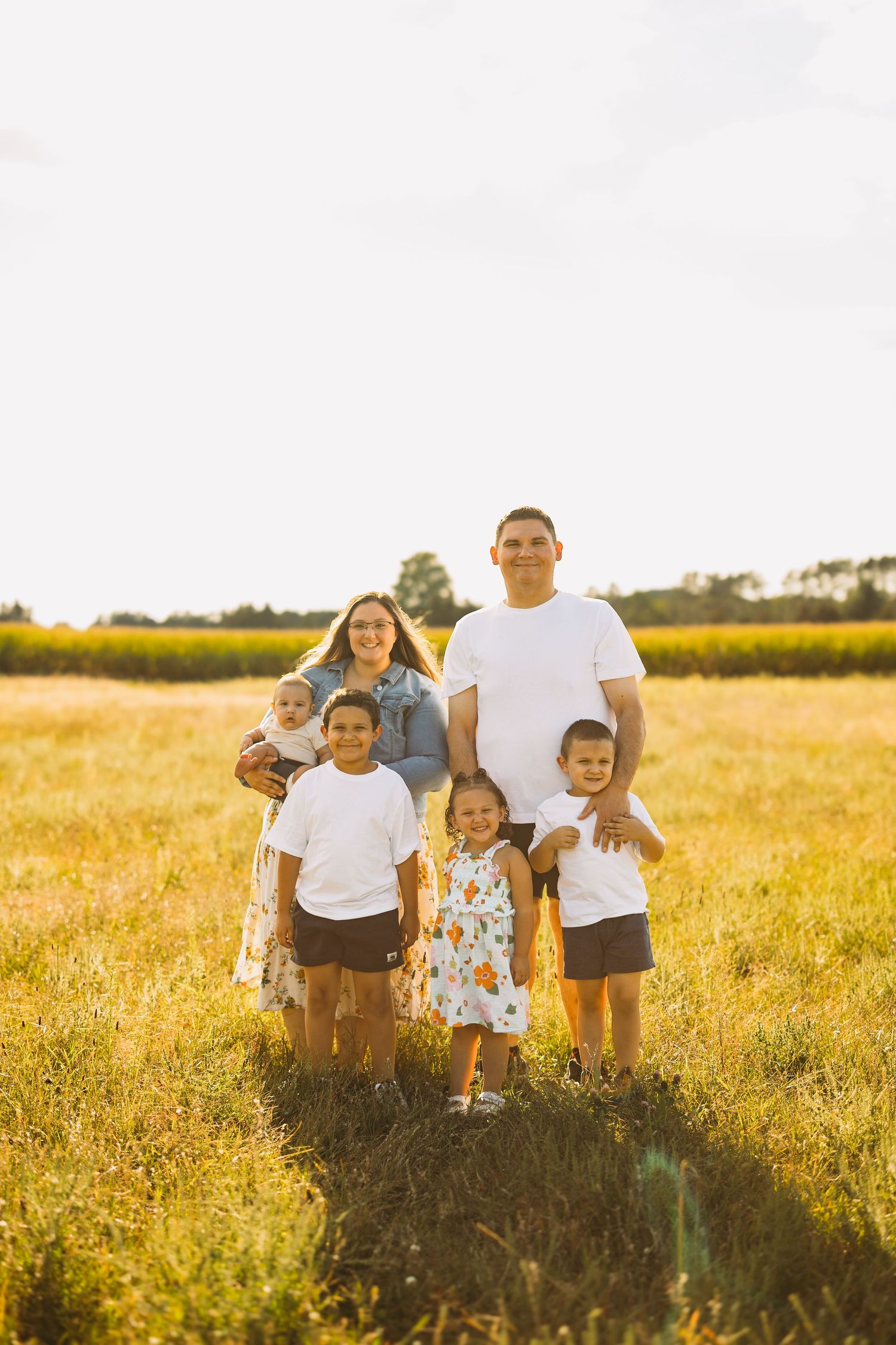 Smiling family of six, with parents, three children and a baby, posing together in a golden grassy field at sunset.