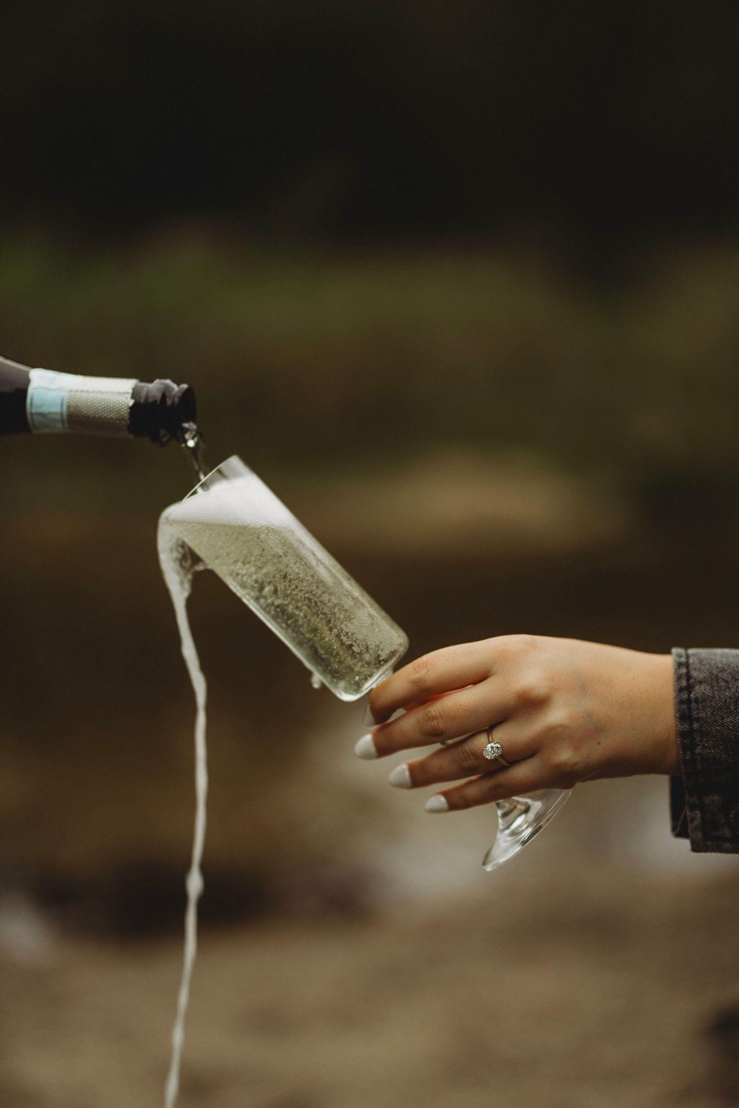 Hand wearing a ring holds a tilted champagne flute as sparkling wine pours from a bottle and foams over the glass outdoors.