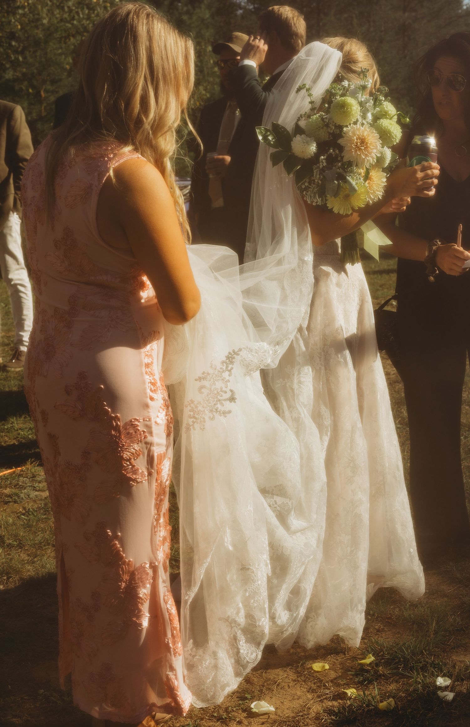 Bride in a lace wedding gown and veil holding a pastel bouquet outdoors while a bridesmaid in a pink lace dress adjusts her train.