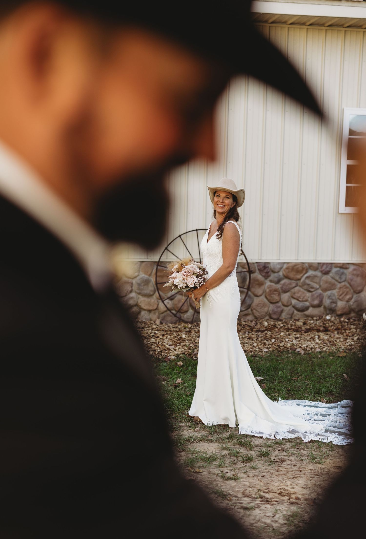 Smiling bride in a white gown and cowboy hat holds a bouquet by a wagon wheel, framed by a blurred groom in the foreground.
