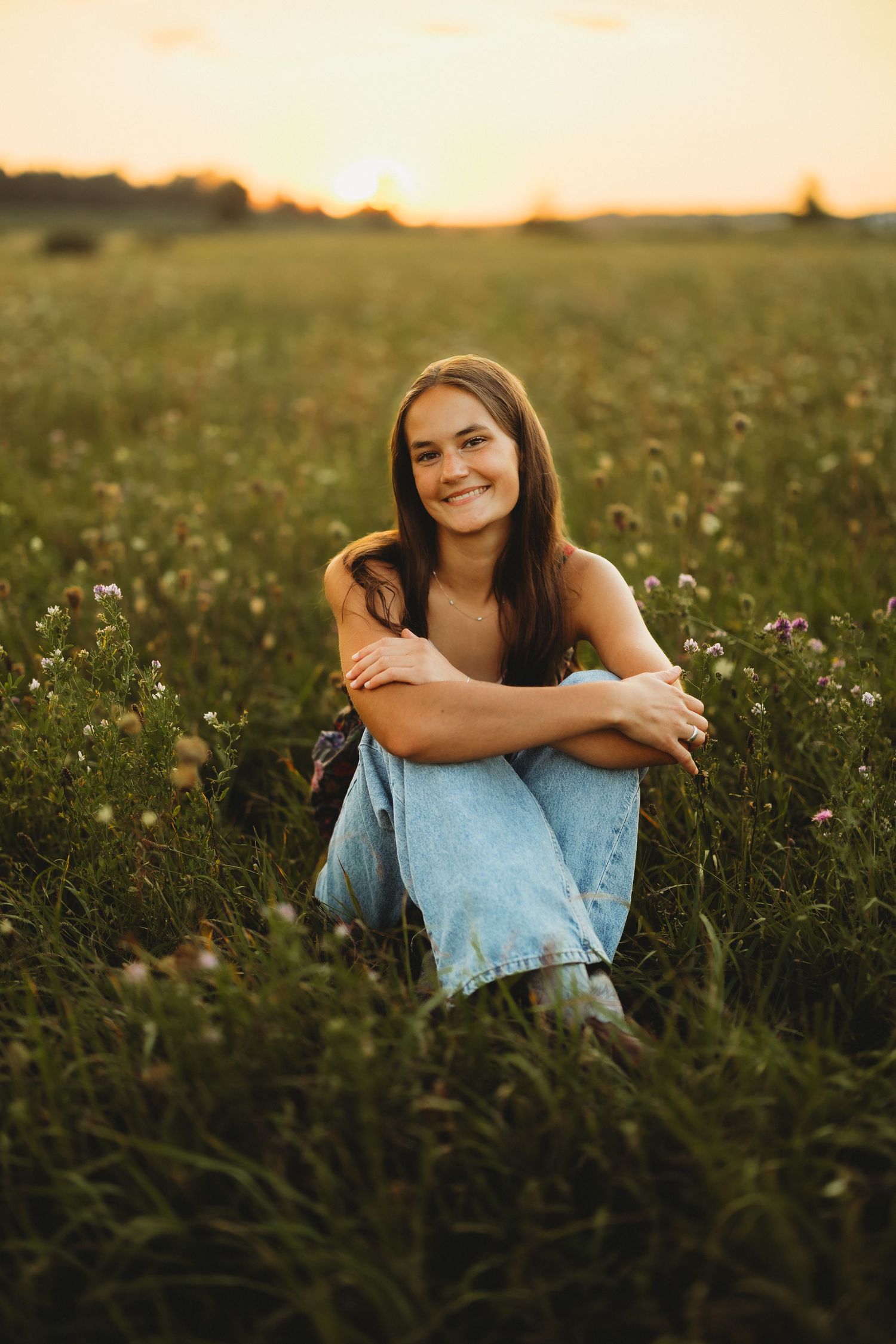 Smiling young woman with long brown hair sits cross-legged in a wildflower field at sunset, wearing light blue jeans and hugging her knees.