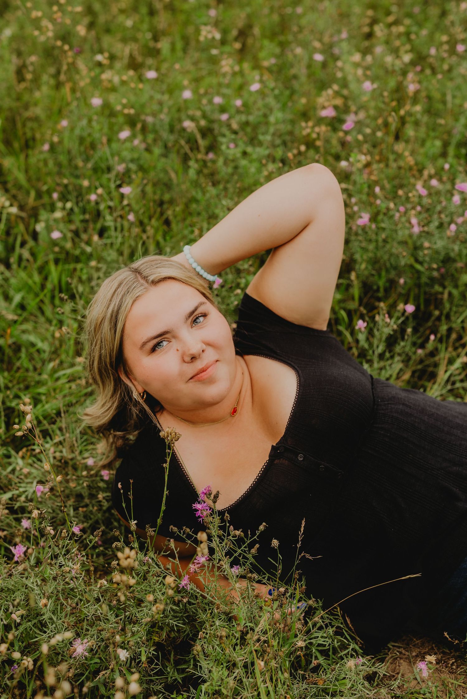 Young blonde woman in black dress lying in wildflower meadow, looking up thoughtfully.