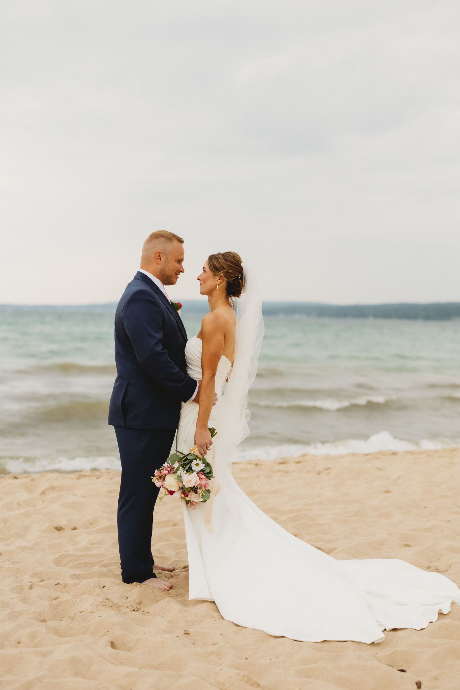 Bride and groom stand together on sandy beach, she in white gown with veil, he in navy suit, ocean waves behind them.