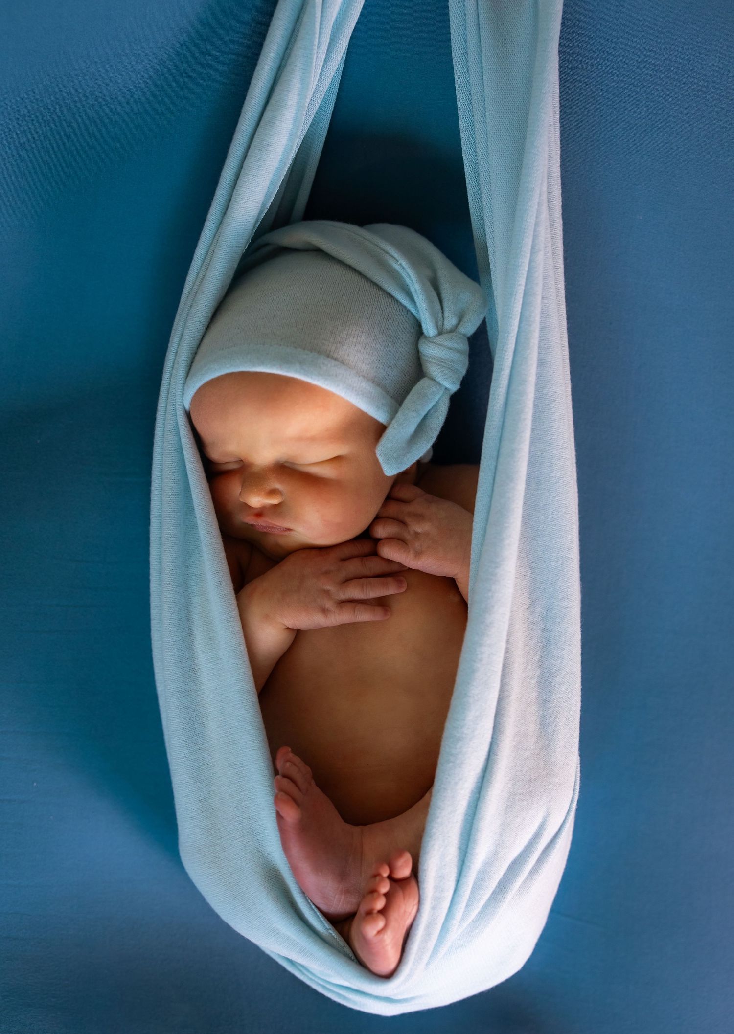 Newborn baby sleeping peacefully in a light blue fabric sling, wearing a matching knotted hat.