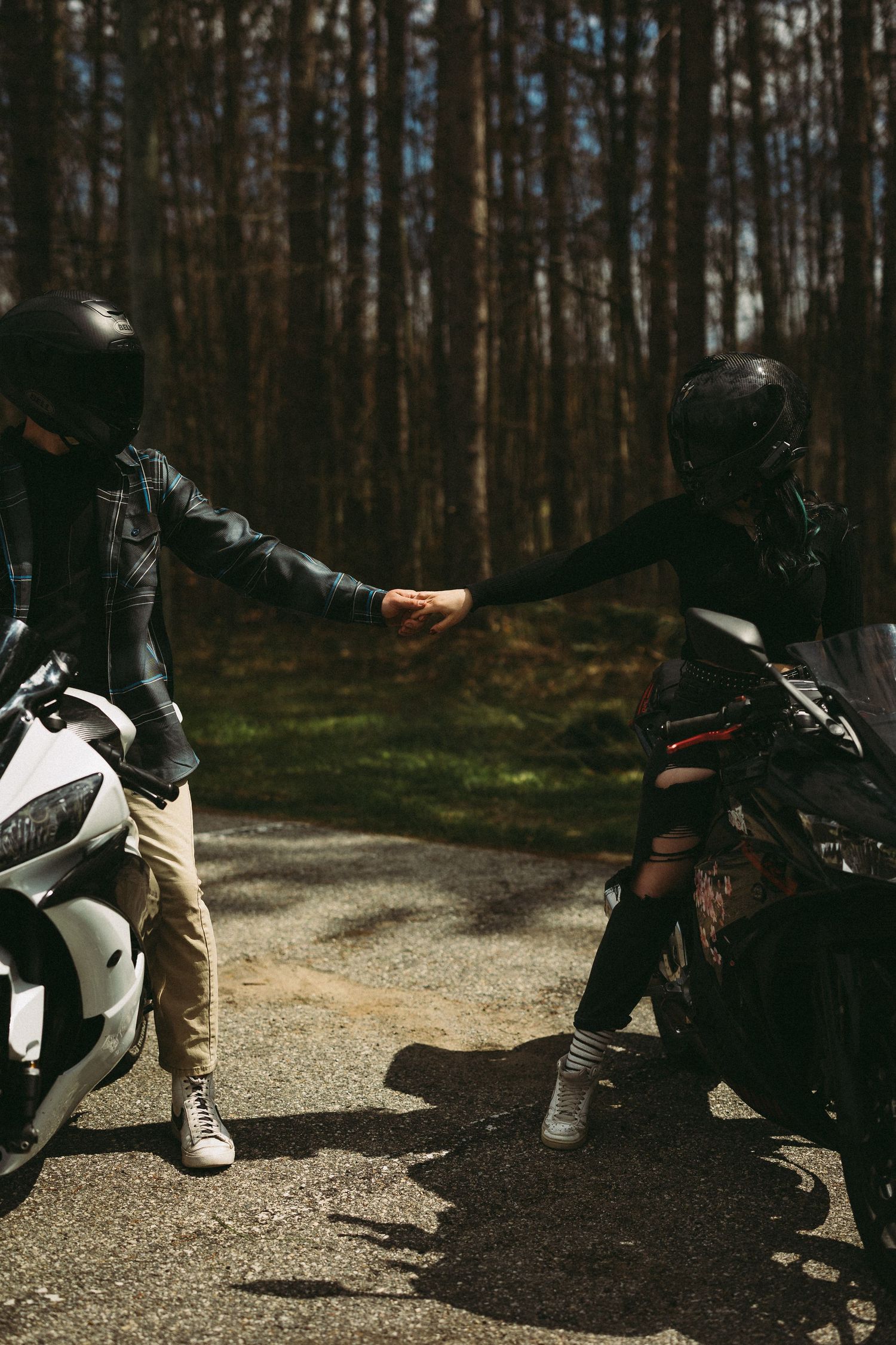 Two motorcyclists in helmets holding hands between their bikes on a forest road on a sunny day.