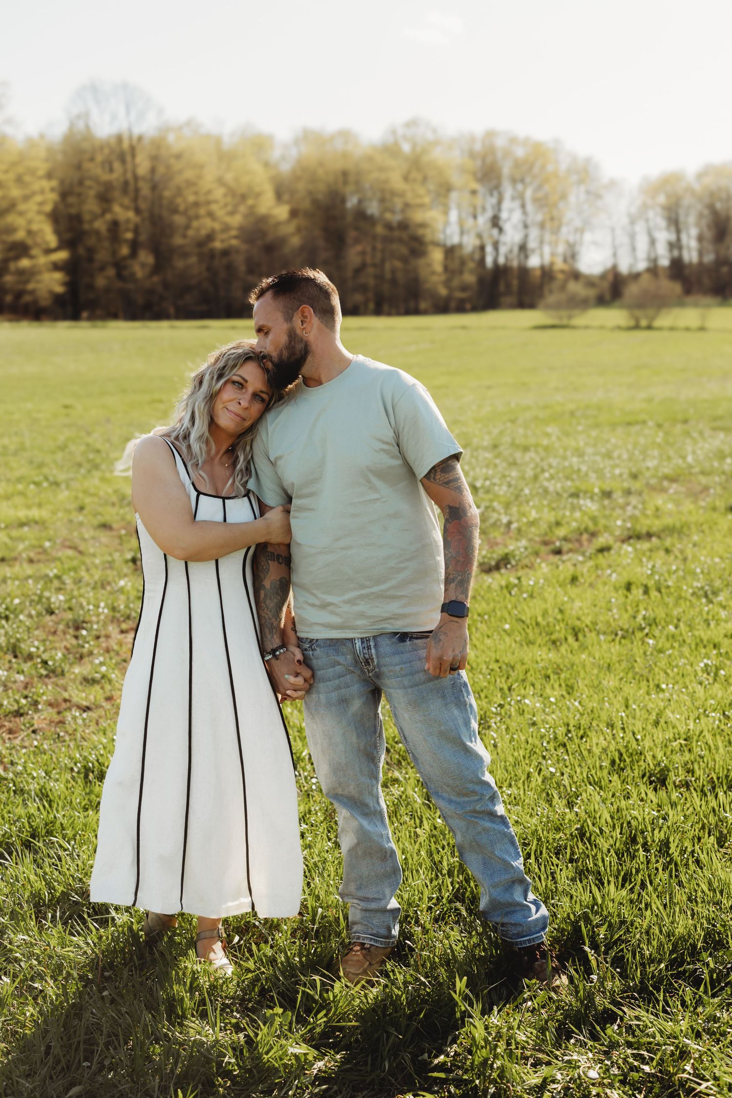 Couple embracing in a sunny green field, woman in white dress leaning on mans shoulder.
