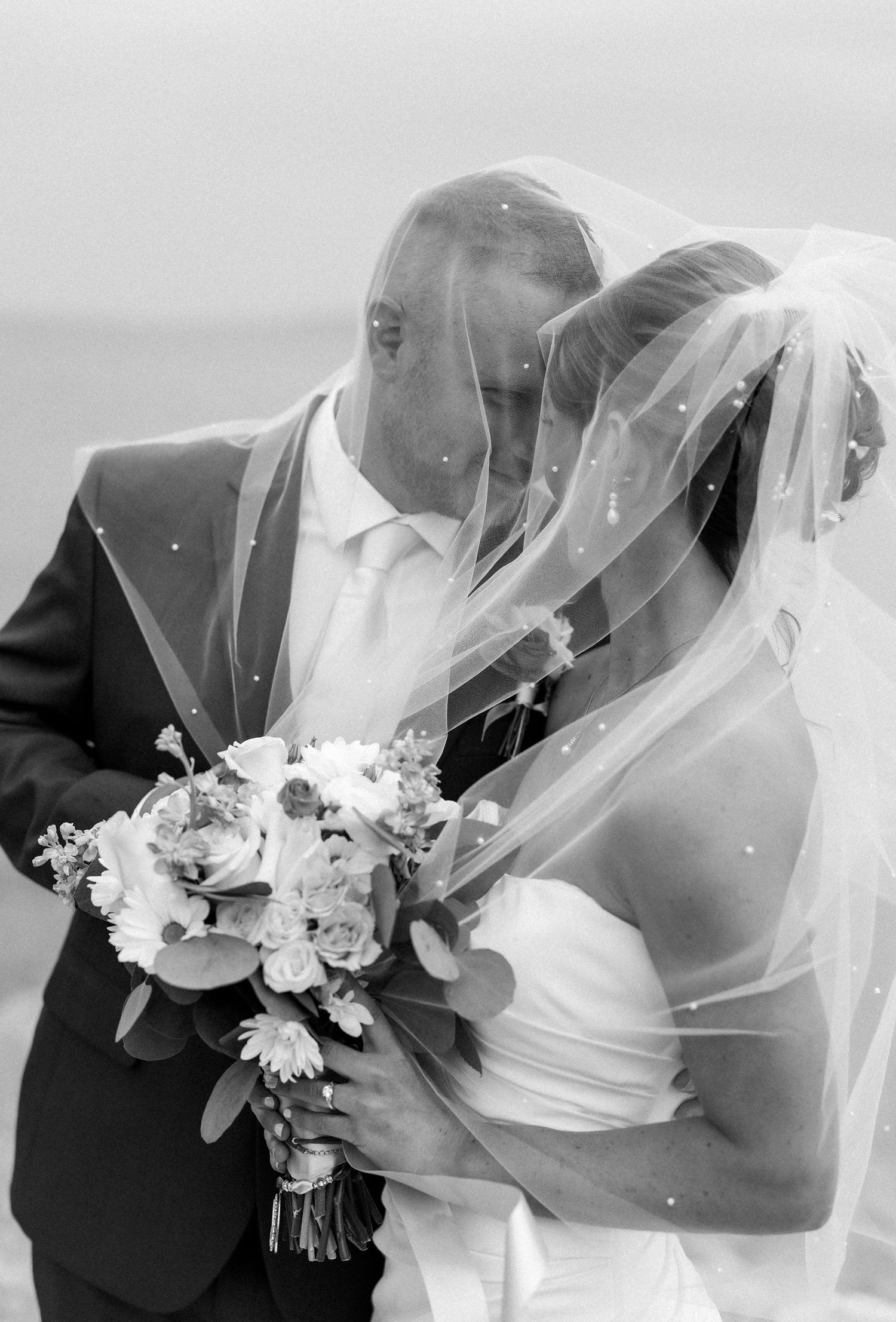 Black and white photo of a bride and groom kissing under a flowing veil, bride holding a bouquet of flowers.