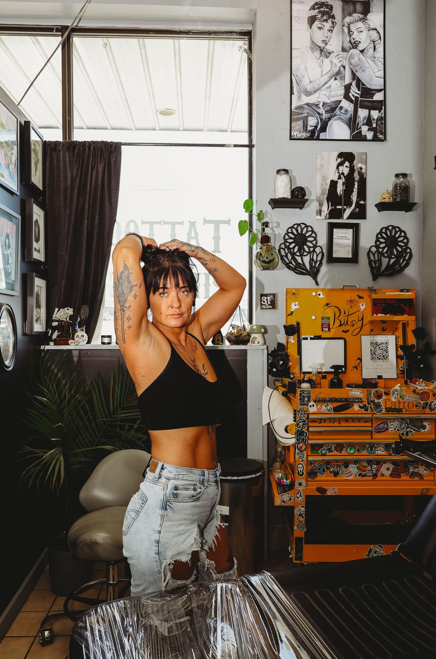 Tattooed woman in black crop top and ripped jeans poses in a tattoo shop with artwork and orange tool chest.