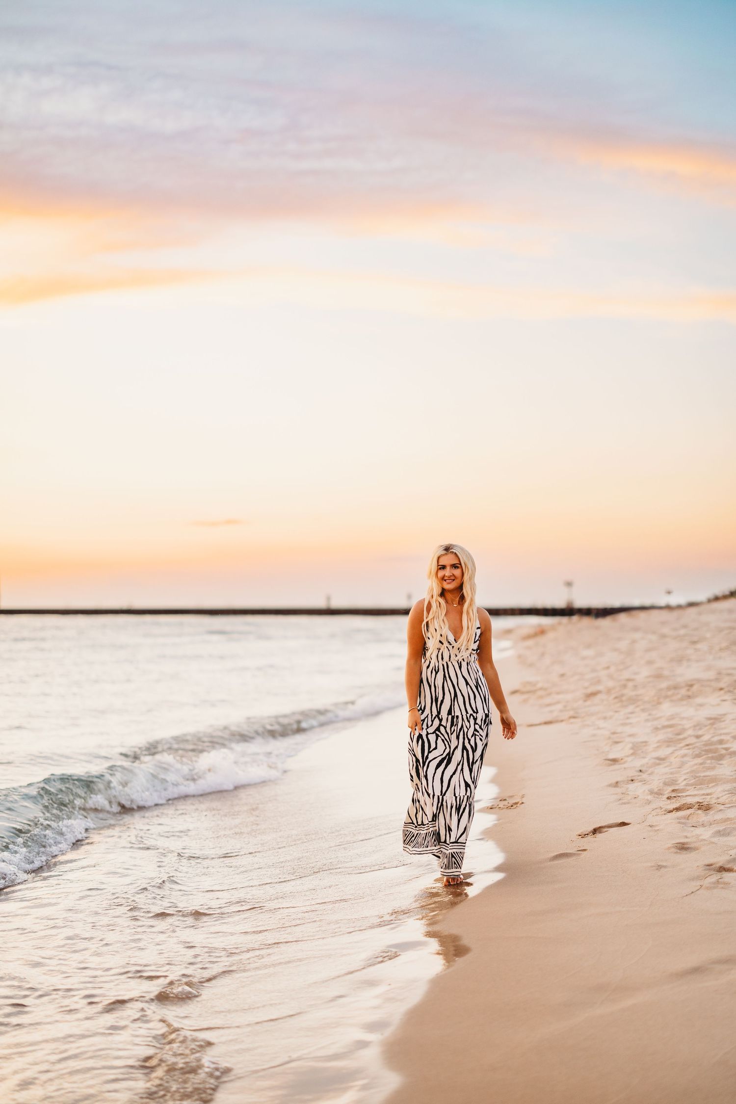 Woman in a zebra-print maxi dress walks barefoot along a sandy beach at sunset with soft waves and a pastel pink sky.