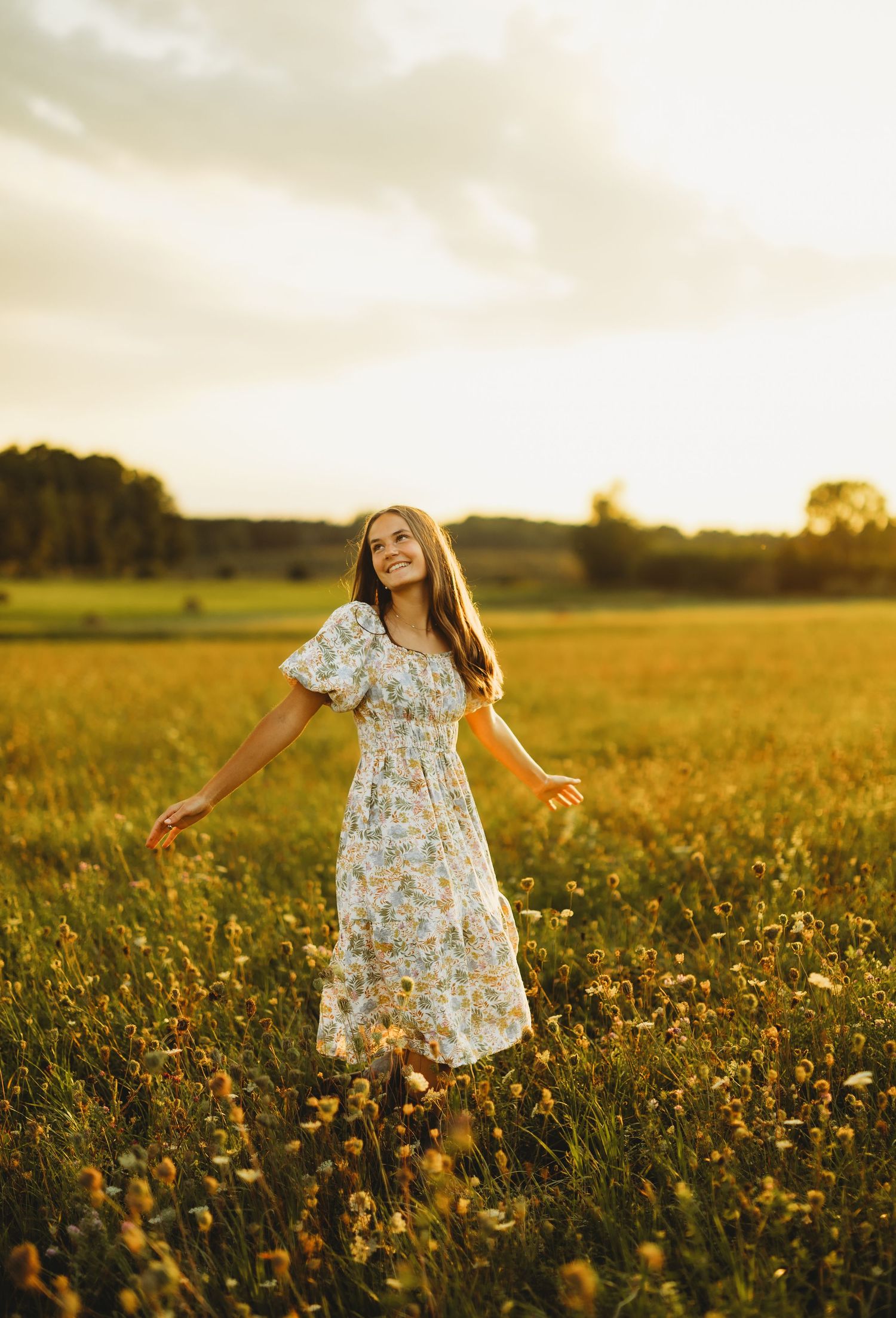 Smiling woman in a floral dress twirls with outstretched arms in a golden wildflower field at sunset.