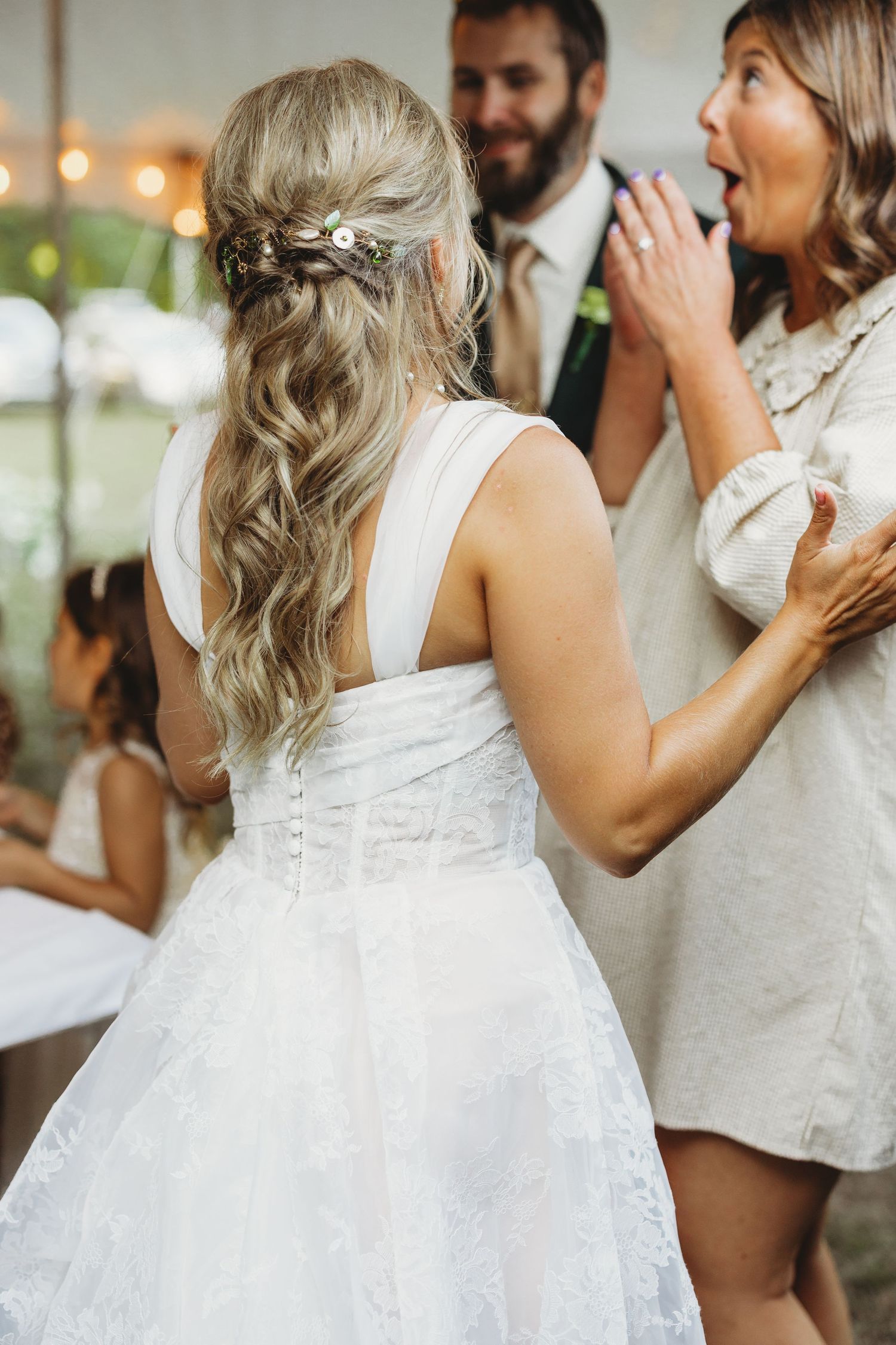 Bride in a white lace wedding dress with a half-up wavy hairstyle and floral hair clip greets a surprised guest at an outdoor reception.