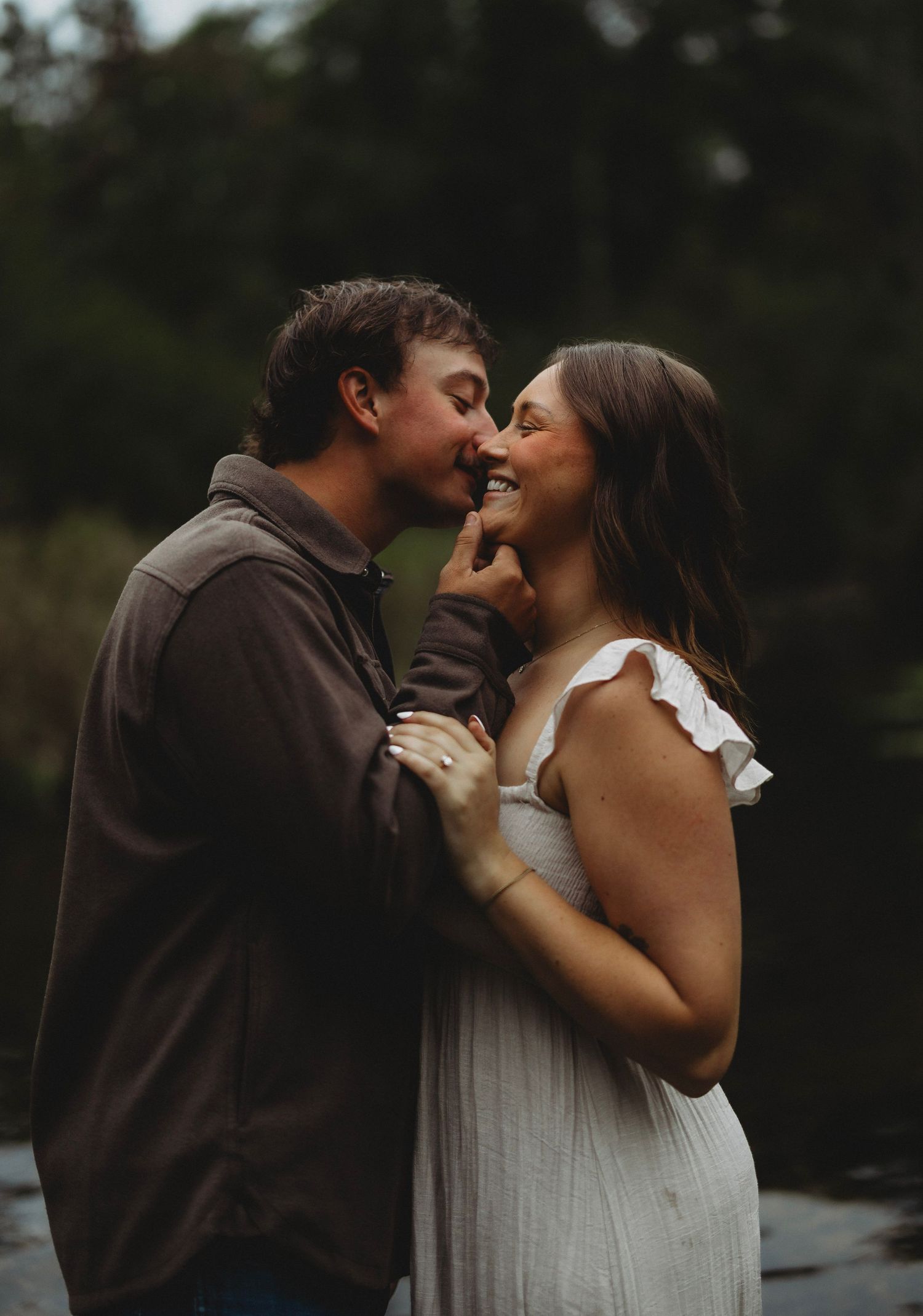 Smiling couple shares an intimate kiss outdoors by a quiet lake, the woman in a white sundress and the man in a brown jacket.