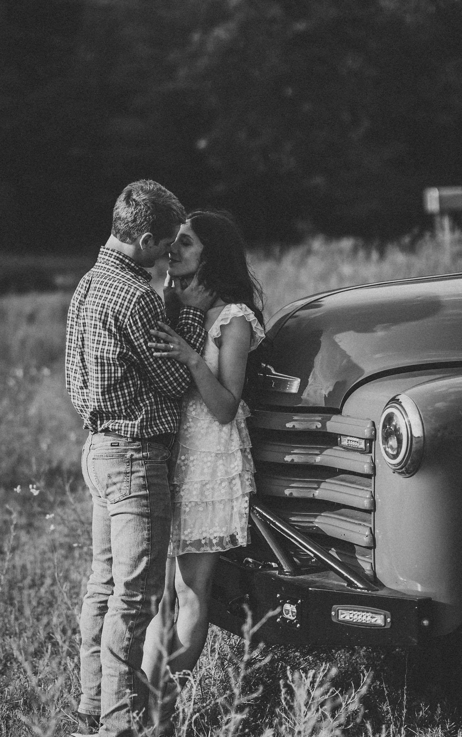 A romantic black and white scene of a couple sharing an intimate moment beside a vintage car.