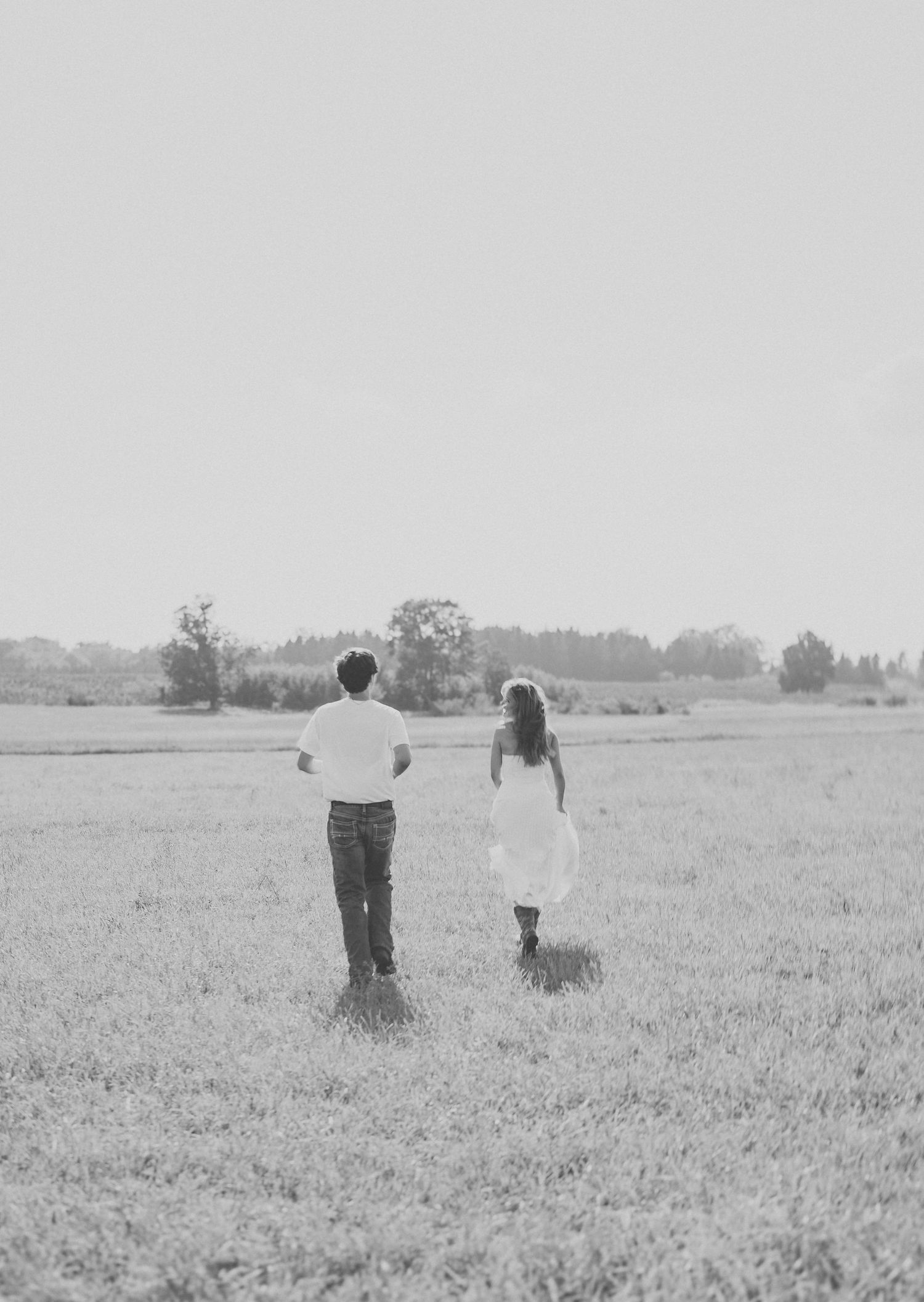 Black and white photo of couple walking through open field, woman in white dress and man in jeans.
