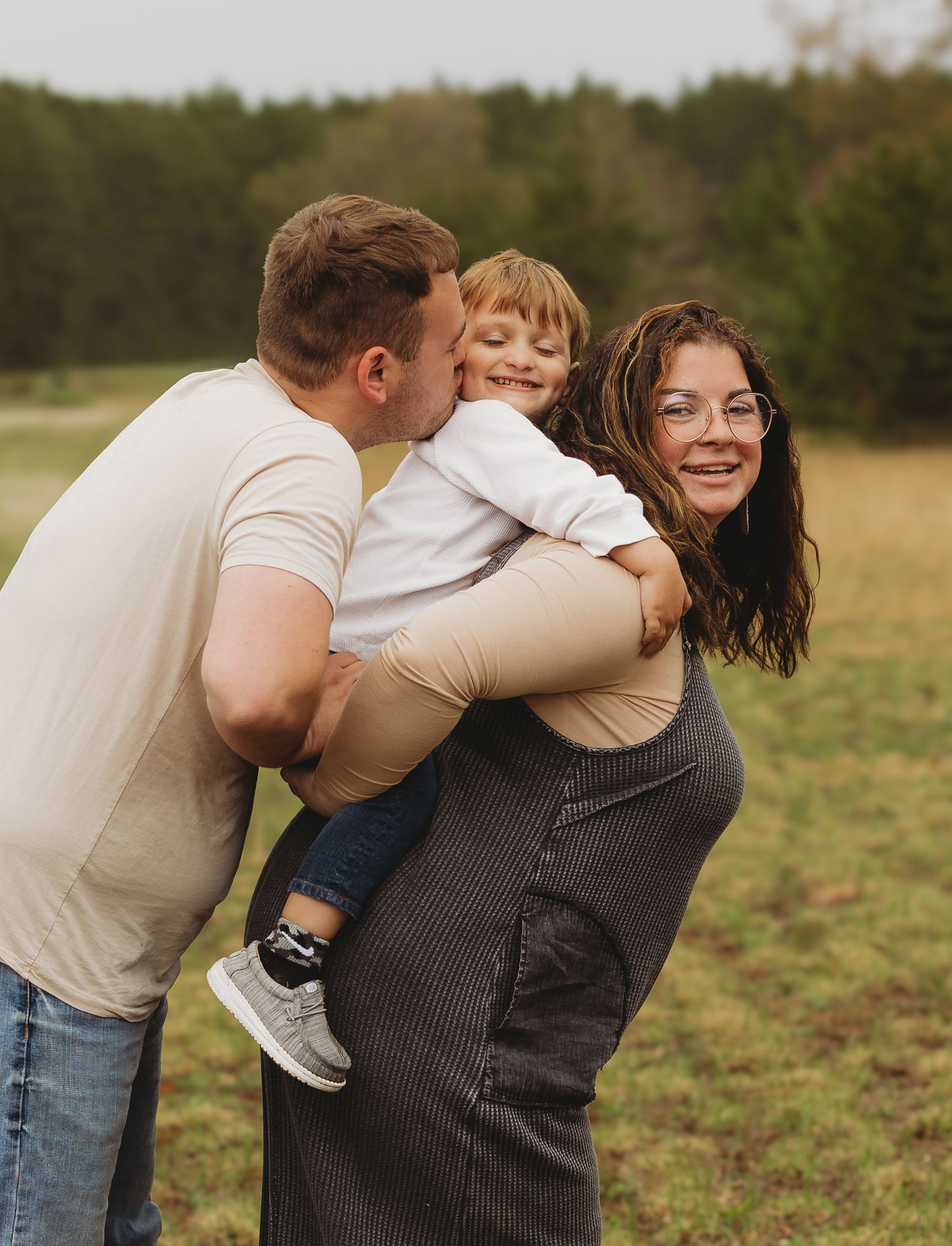 A happy family of three poses outdoors, with a woman giving a child a piggyback ride while a man kisses the child.