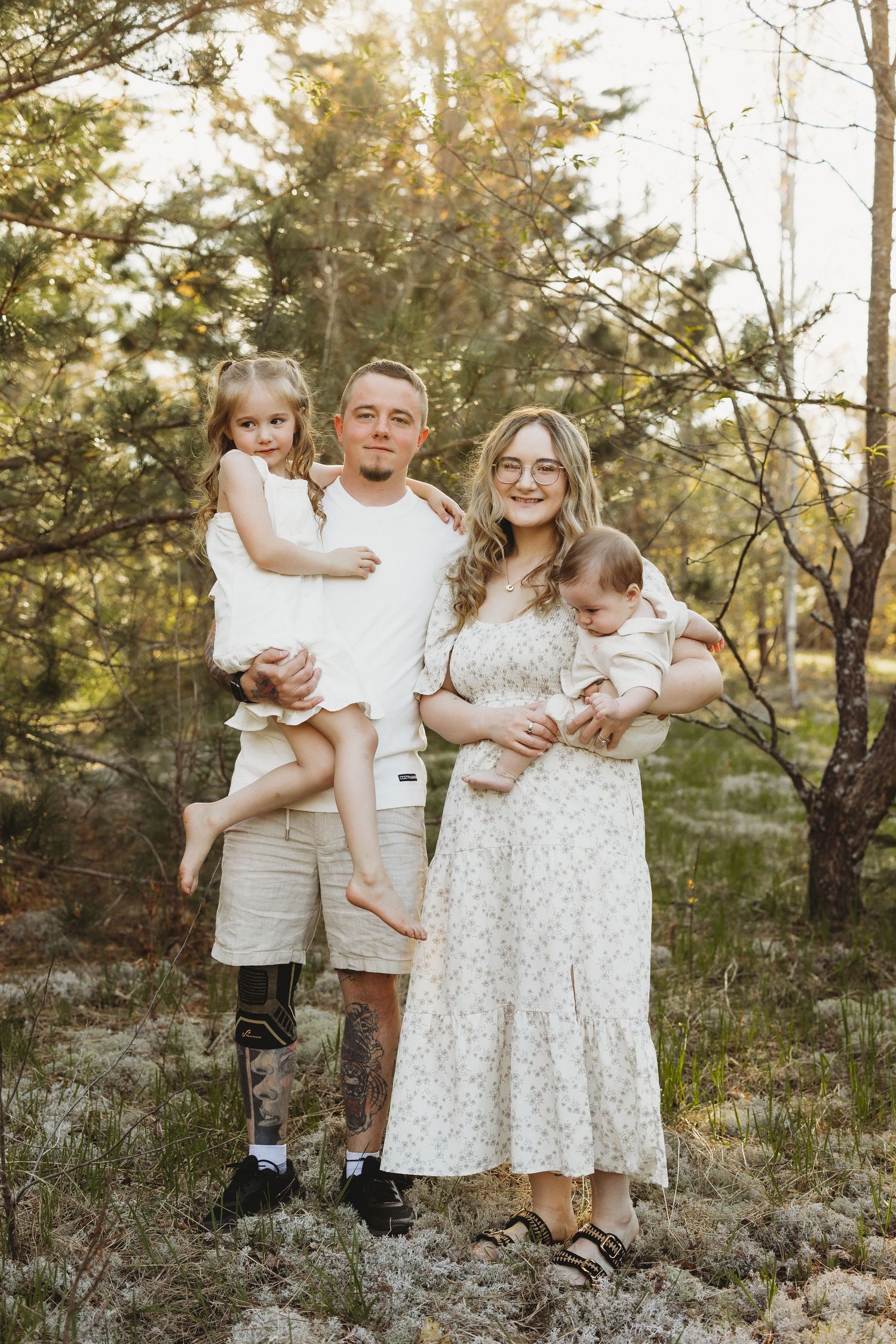 Young family standing in sunlit forest, man holding girl, woman cradling baby, all wearing white and cream outfits.