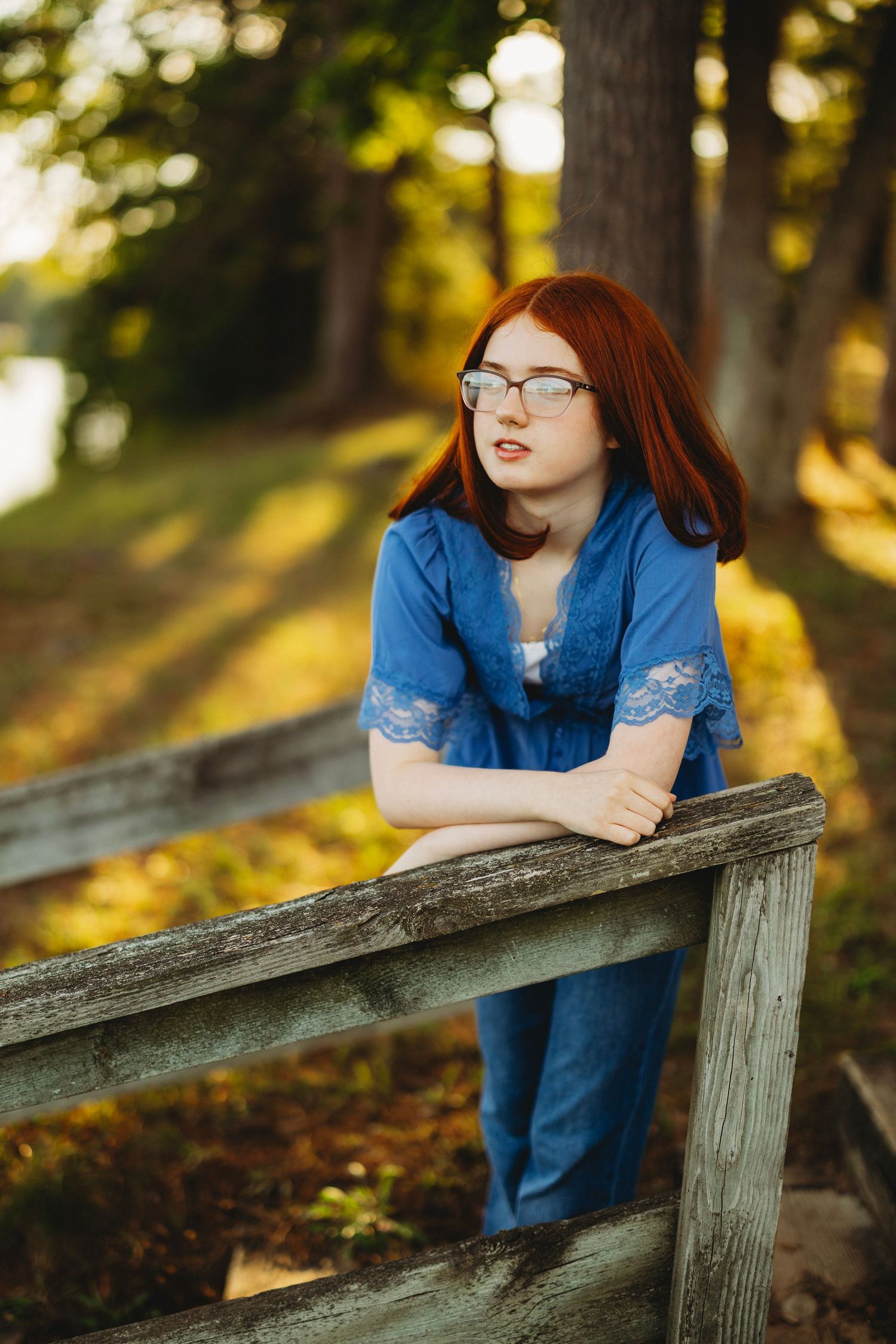 Red-haired girl with glasses in a blue top and jeans leans on a weathered wooden railing in a sunlit, tree-filled park.