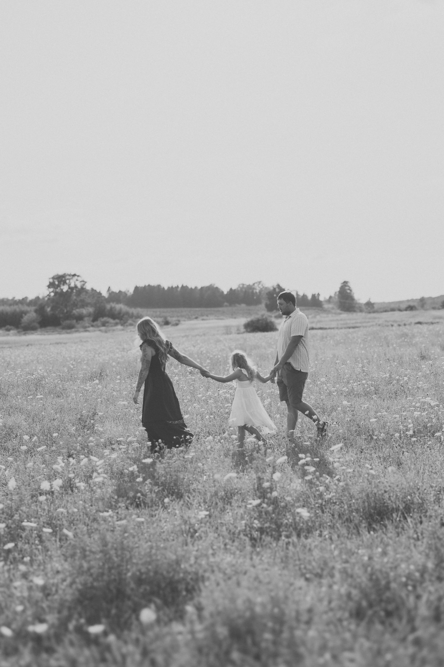 Black and white photo of a family of three holding hands and walking through a wildflower meadow at sunset.
