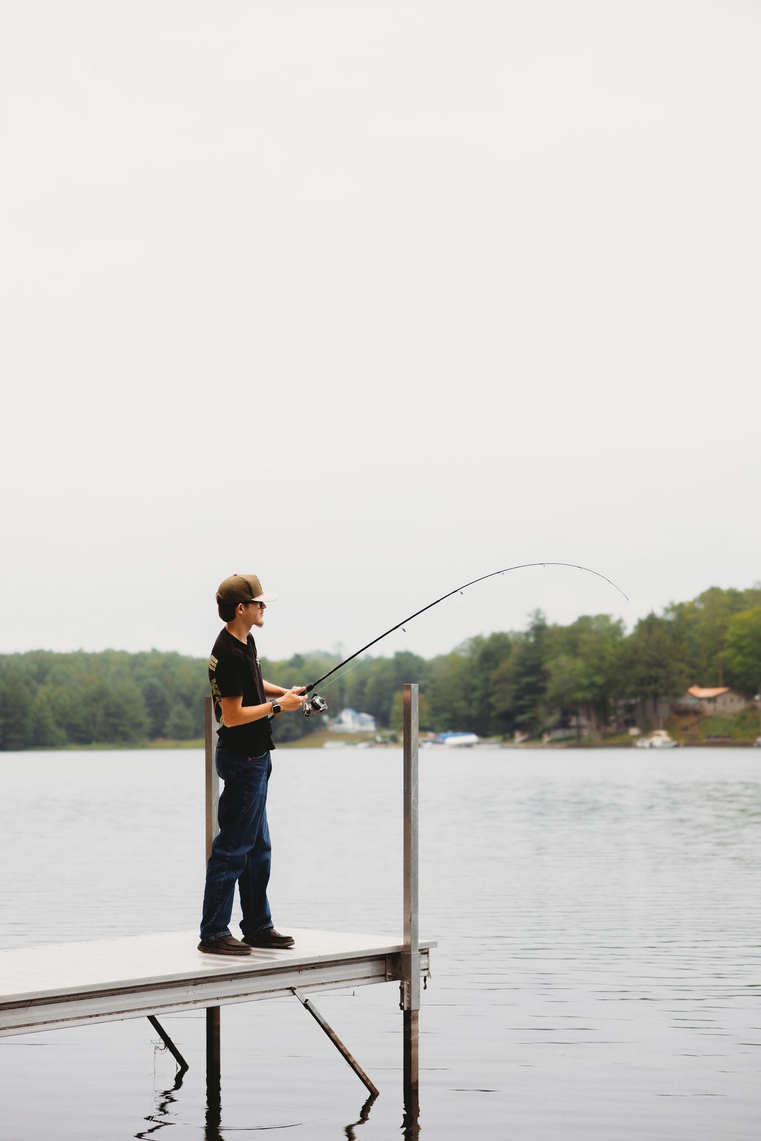 Young man in a dark shirt and jeans fishing with a rod from a wooden dock on a calm lake with a tree-lined shore.