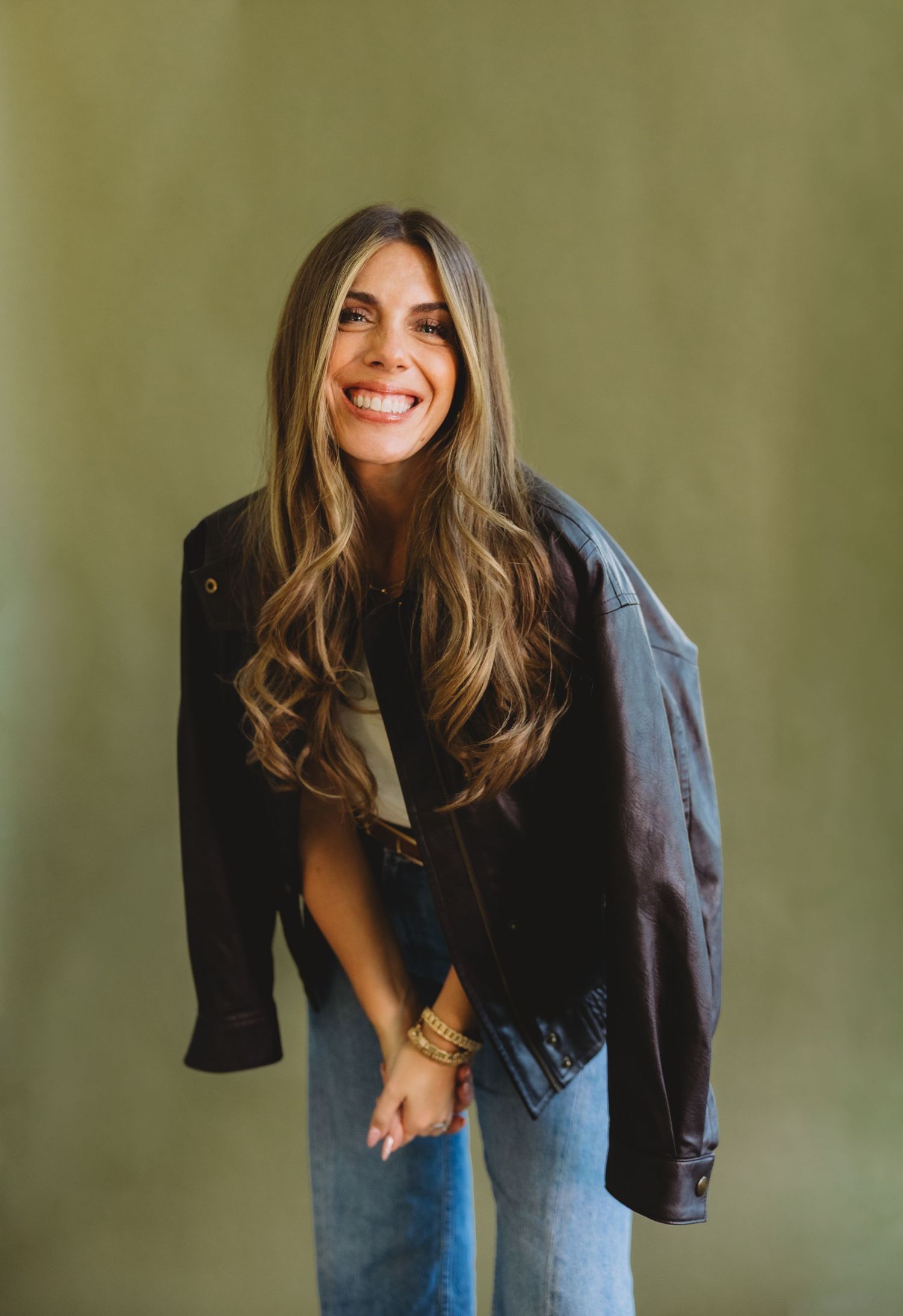 Smiling woman with long wavy hair leans forward in a dark leather jacket and blue jeans against an olive green backdrop.