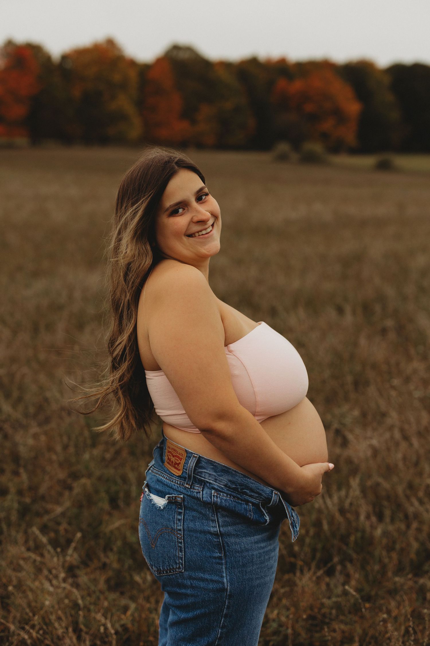 A person in a white top and jeans poses outdoors in a fall field at sunset with autumn colors in the background.
