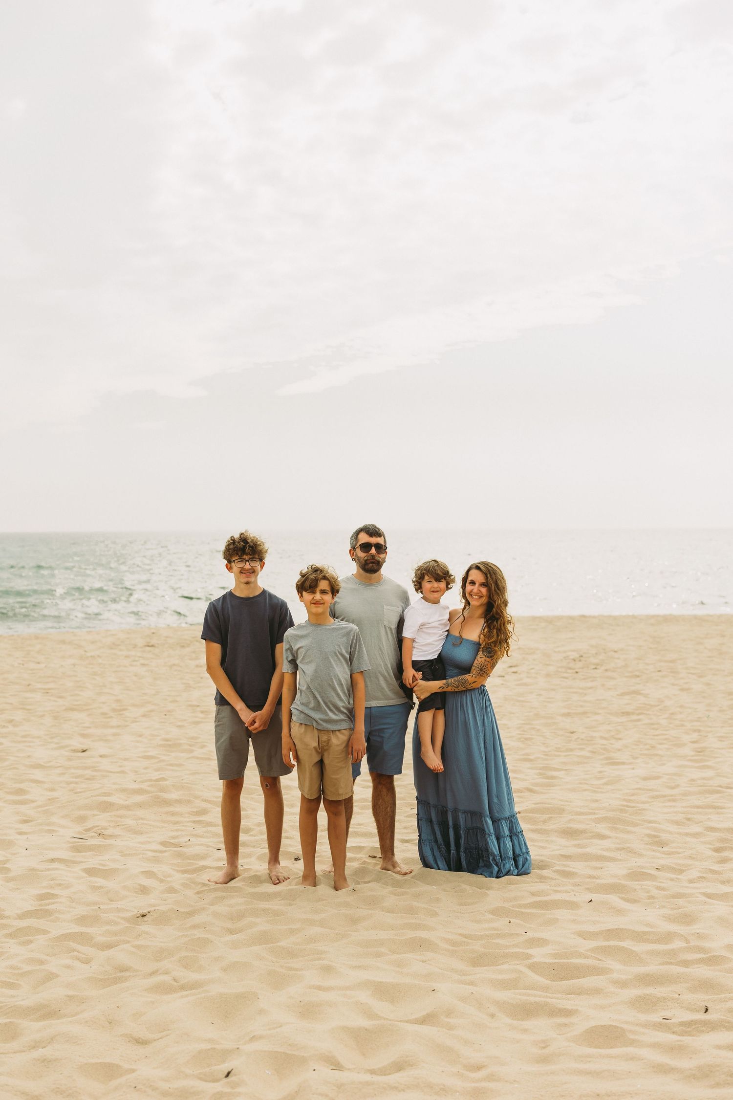 A family of five poses together on a sandy beach with the ocean in the background.