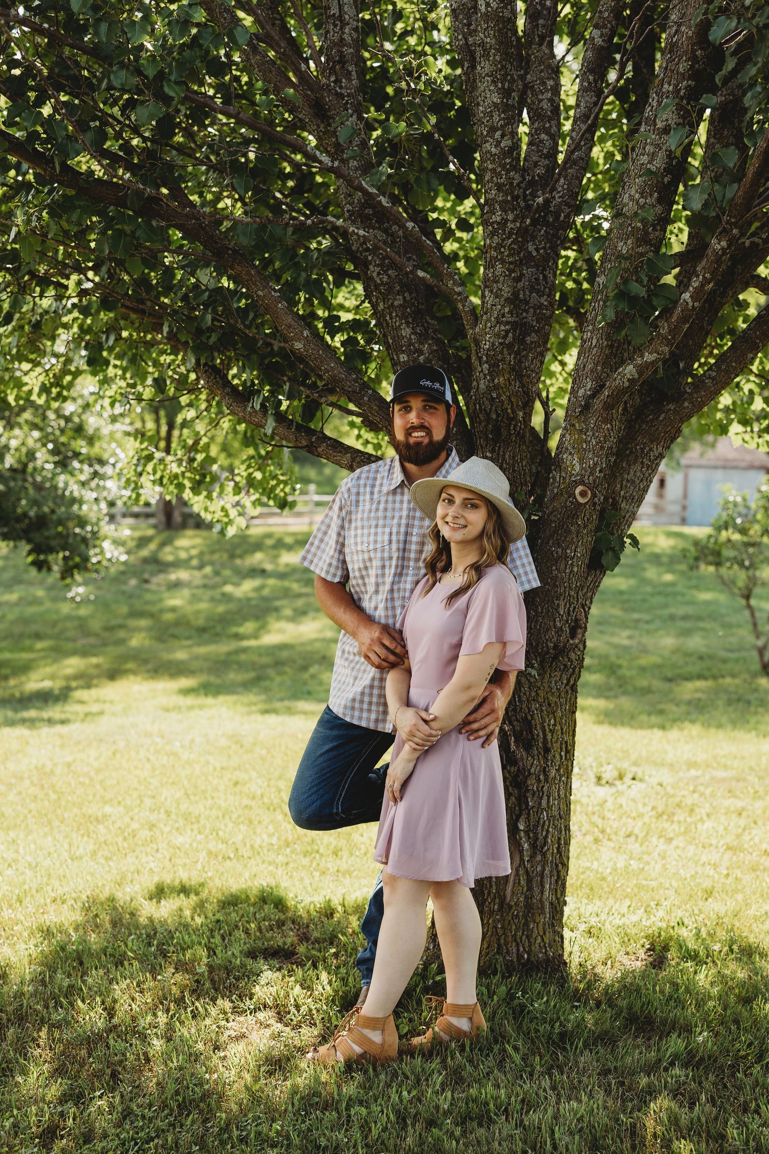 A couple embraces beneath a shady tree in a sunny park setting on a warm summer day.