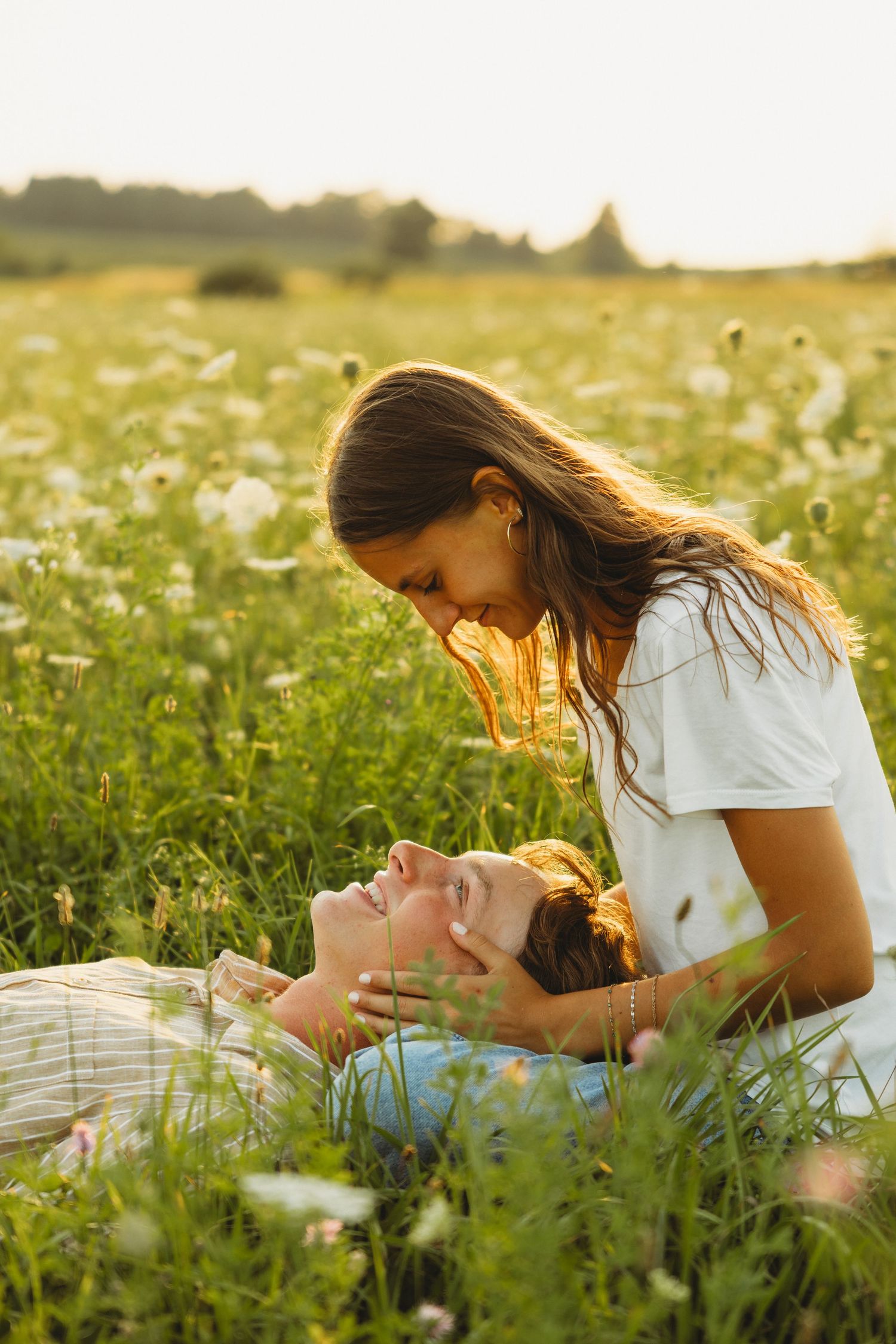A couple shares a tender moment in a sunlit wildflower field, woman leaning down to kiss man lying in grass.