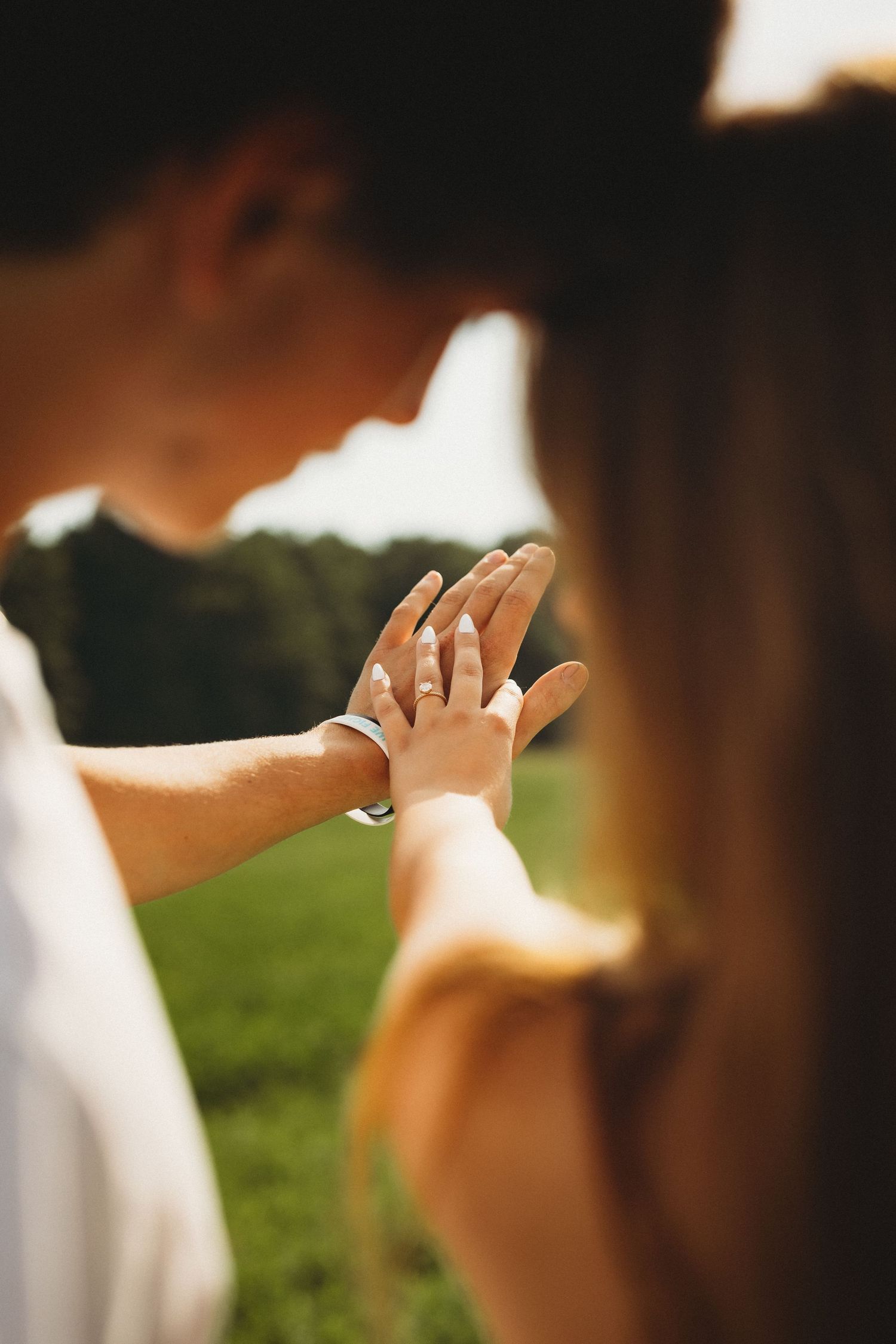 Couple holding hands outdoors in golden sunlight, showcasing an engagement ring on womans finger in a romantic field setting.