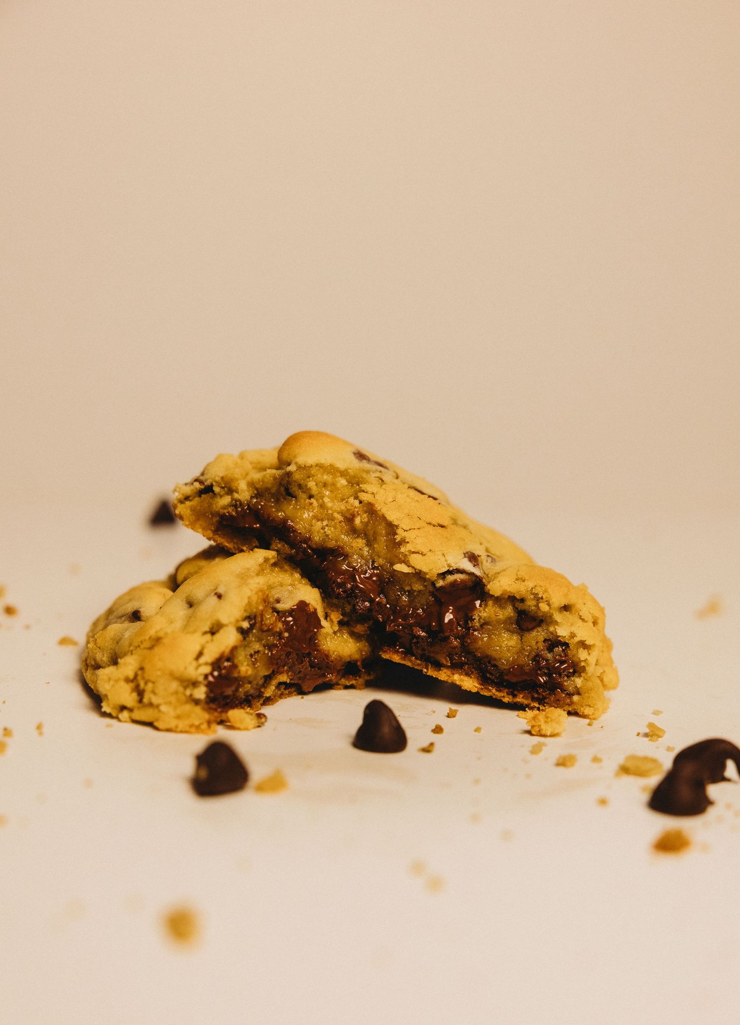 A freshly baked chocolate chip cookie broken in half revealing gooey melted chocolate, with crumbs scattered on white surface.